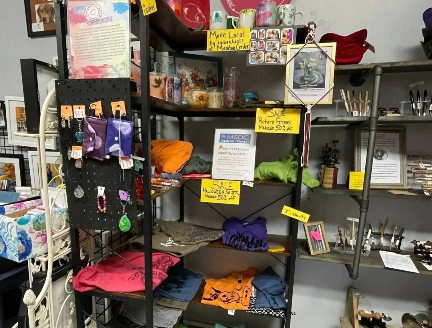 Shelves displaying picture frames, Halloween shirts, T-shirts, mugs, and jewelry at a thrift store with sale signs and promotional materials.