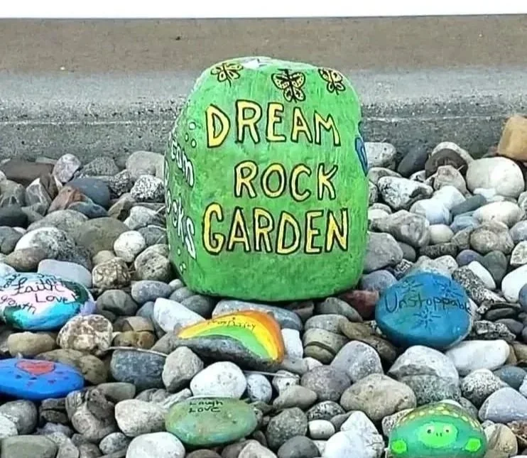 Colorfully painted rocks with messages and designs, centered around a green rock with yellow lettering that reads "Dream Rock Garden" surrounded by other decorated rocks on a bed of small stones.