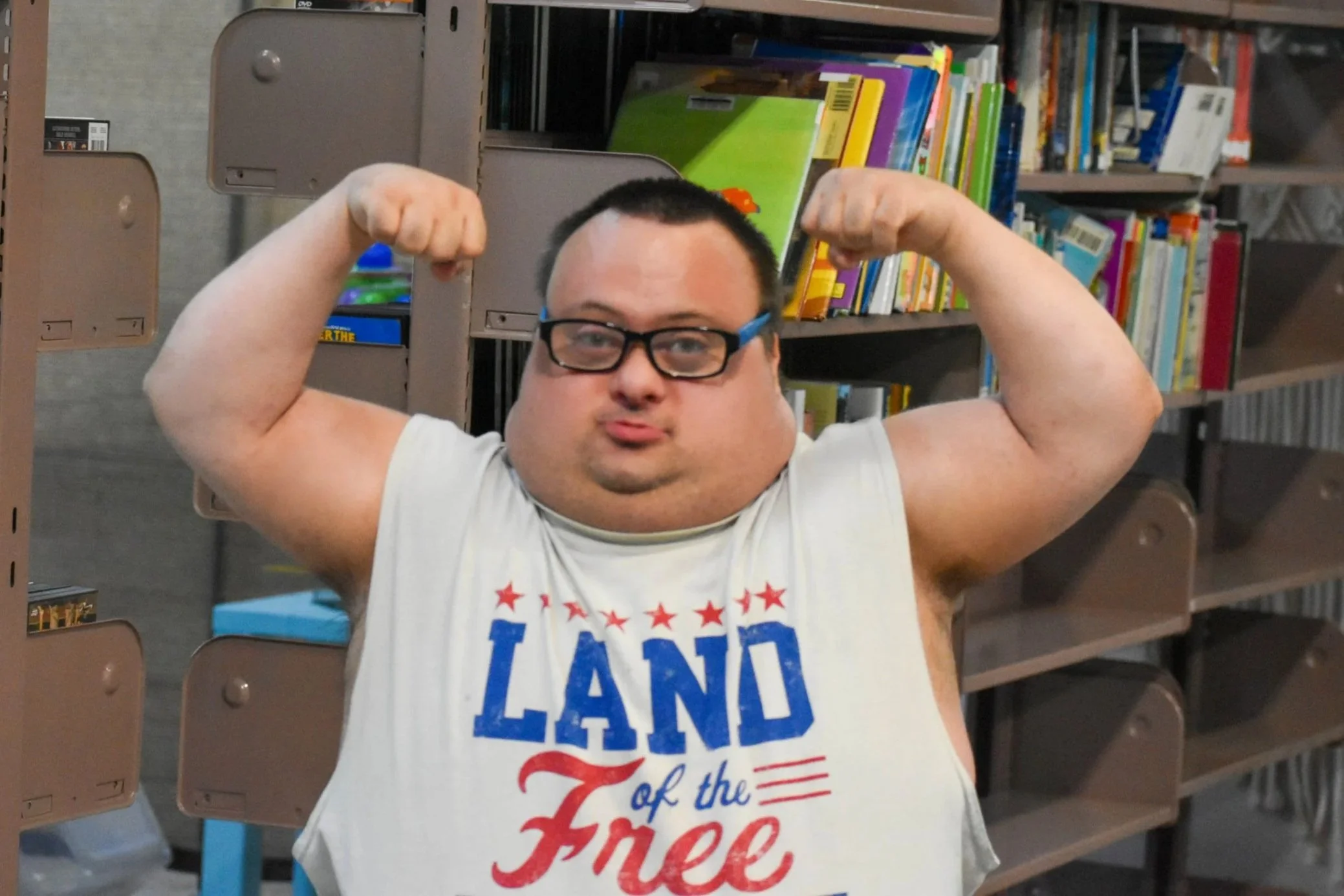 A man wearing glasses and a sleeveless shirt with 'Land of the Free' printed on it, flexing his arms in a library or bookstore with shelves of books in the background.