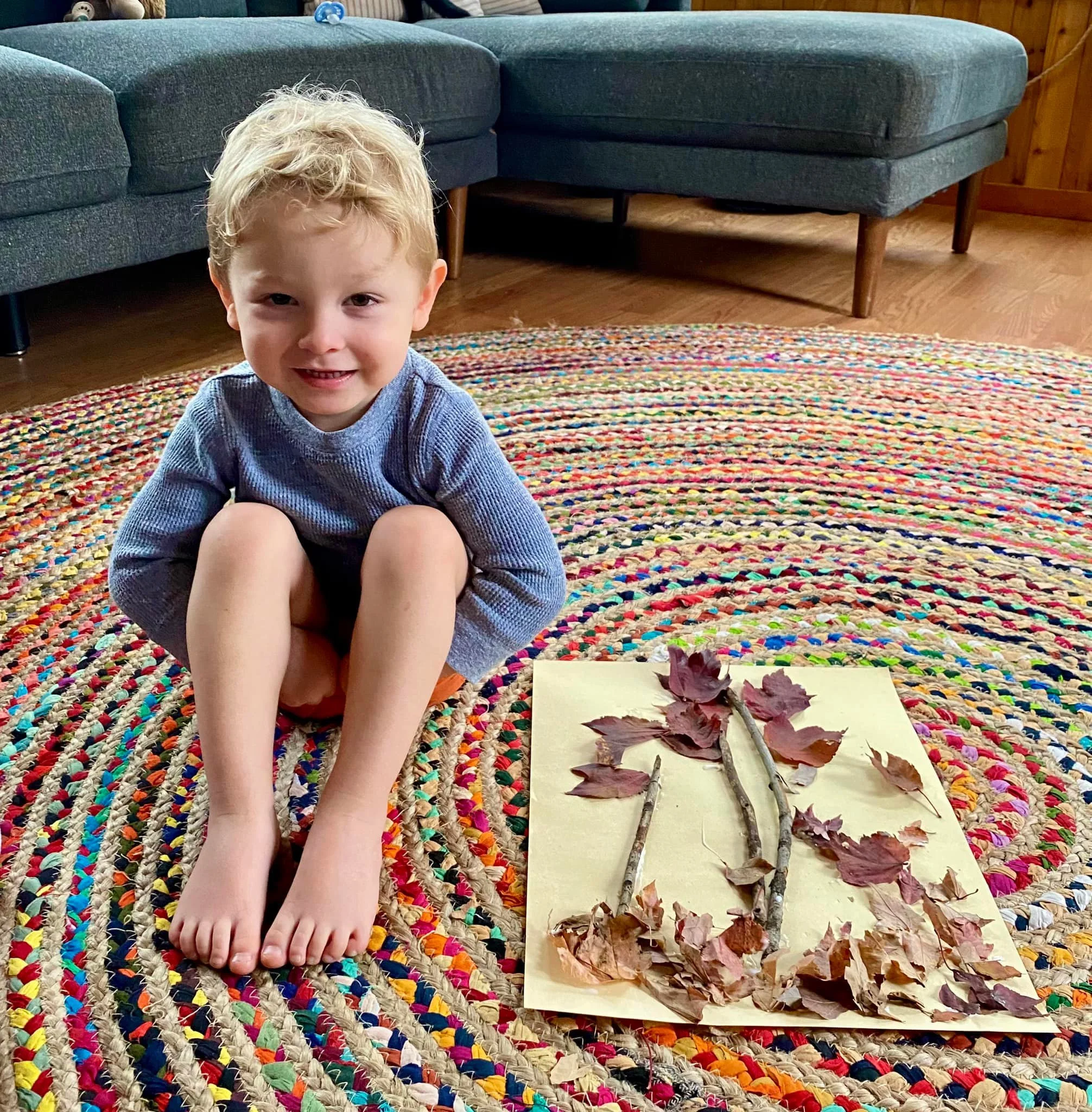 A young boy with blonde curly hair, wearing a blue long-sleeve shirt, sitting on a colorful braided rug indoors. In front of him is a paper with sticks and fallen leaves arranged on it, likely a craft project. The background shows a gray sectional sofa and wooden wall paneling.