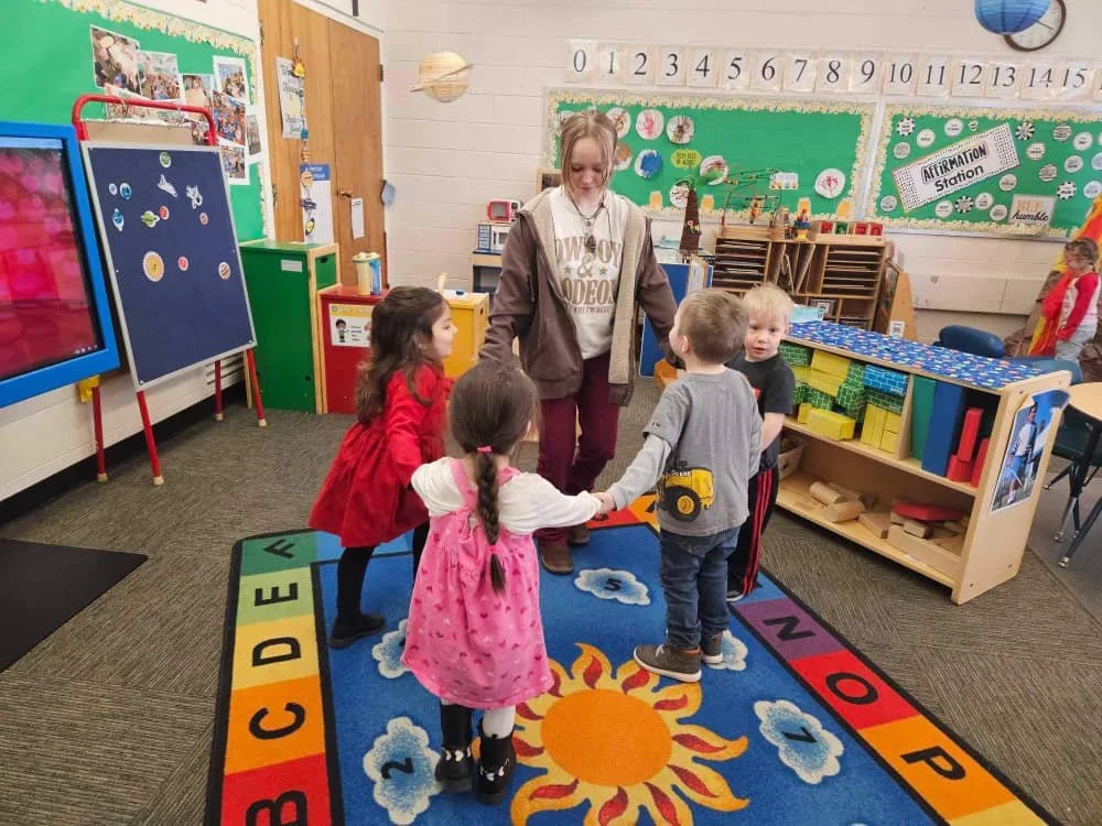 A teacher and four young children standing on a colorful educational rug in a classroom, holding hands in a circle.