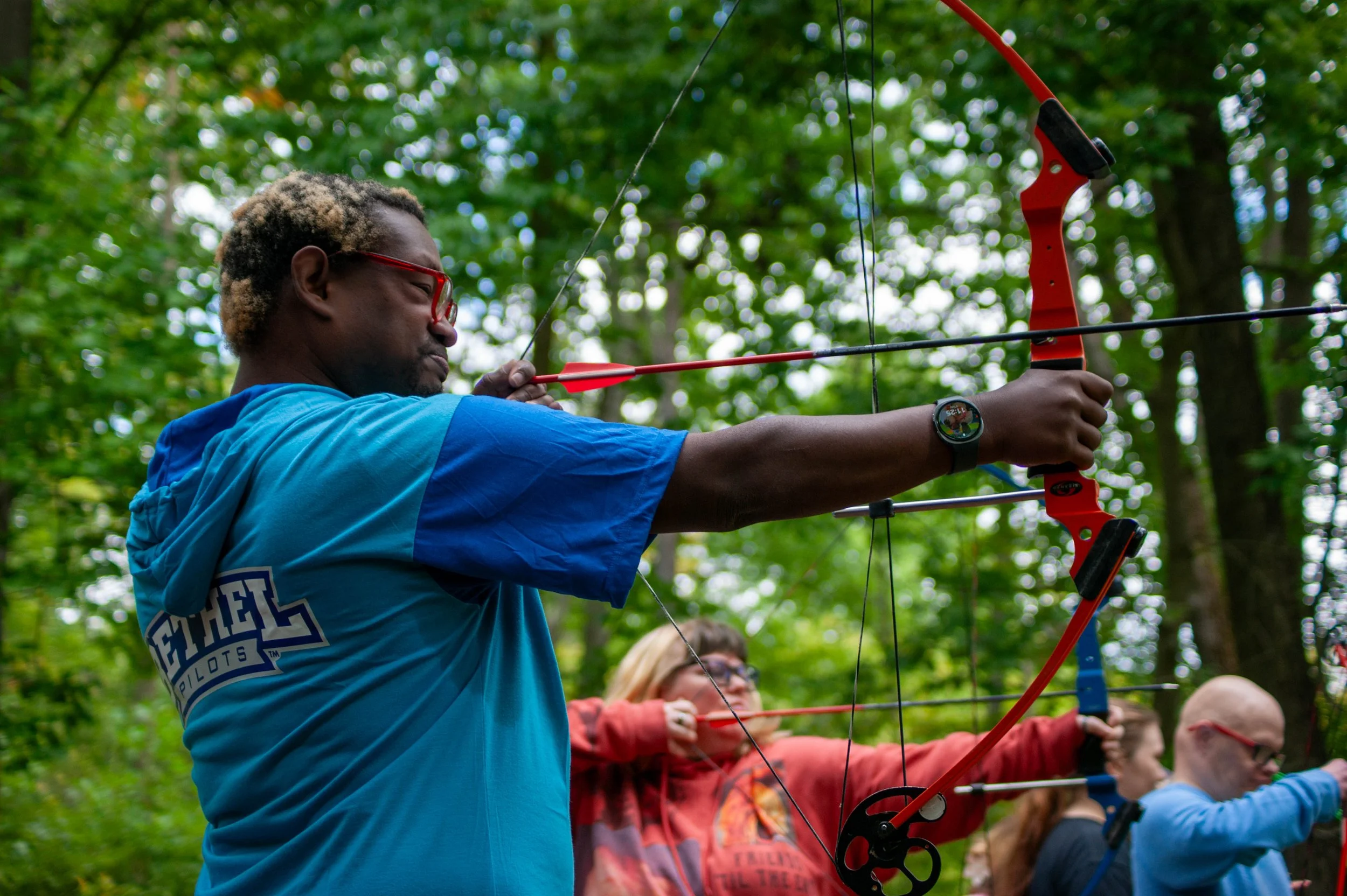 A man aiming a red and black bow at a target outdoors among trees, with other people in the background also holding bows.