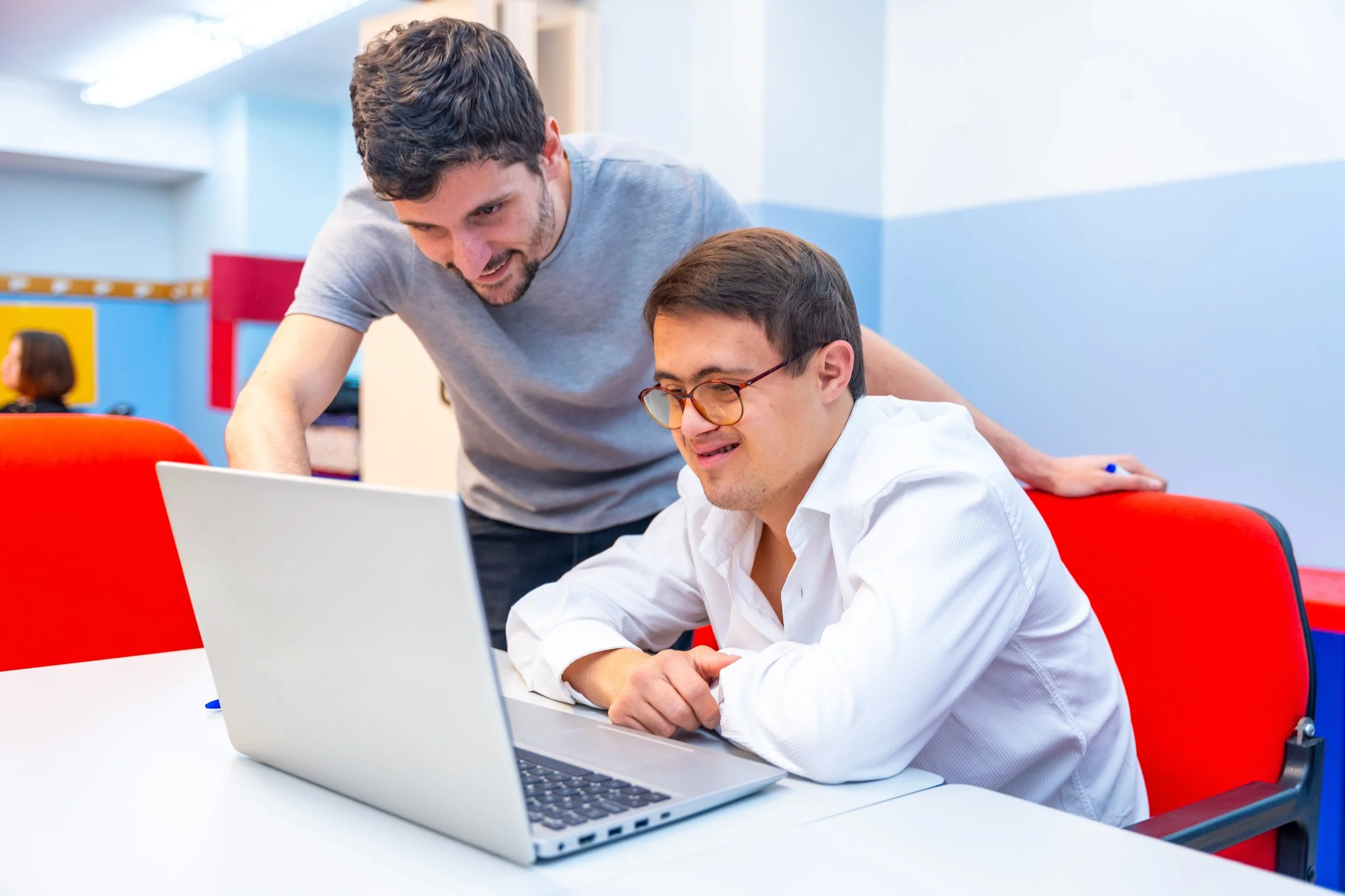 Two men look at a laptop screen in a brightly lit room with blue and yellow walls. One man is sitting and wearing glasses, while the other is standing and leaning in. Both appear focused and engaged.
