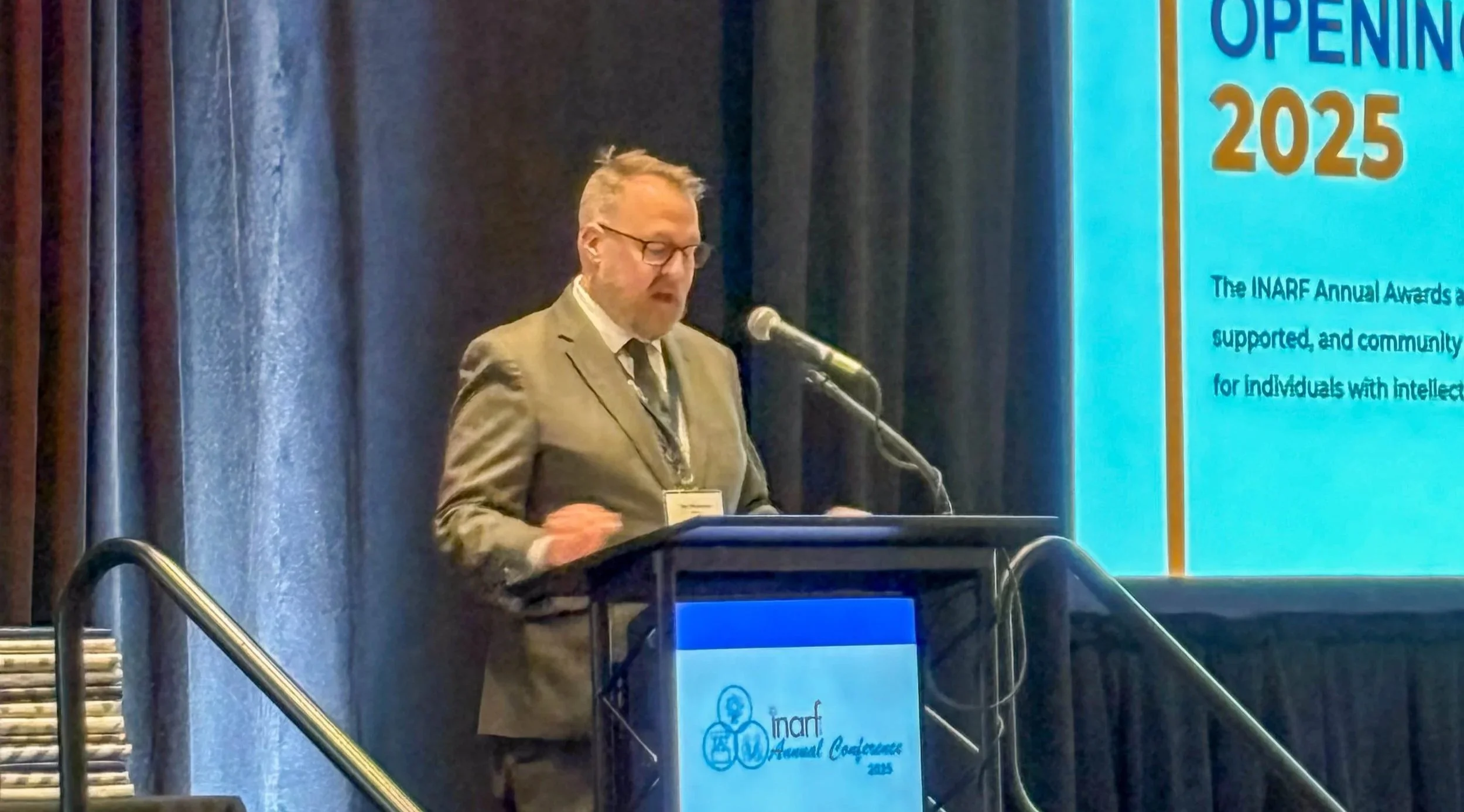 A man in a gray suit, glasses, and a patterned tie stands at a podium with a laptop, speaking at a conference. Behind him is a large screen displaying text about the INARF Annual Awards, with the words "Opening 2025" visible.