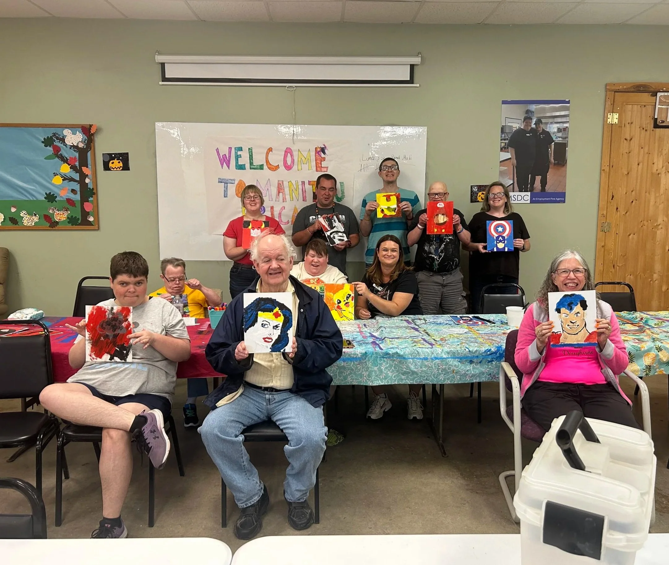 Group of people at an art activity holding up painted portrait artworks inside a room with a