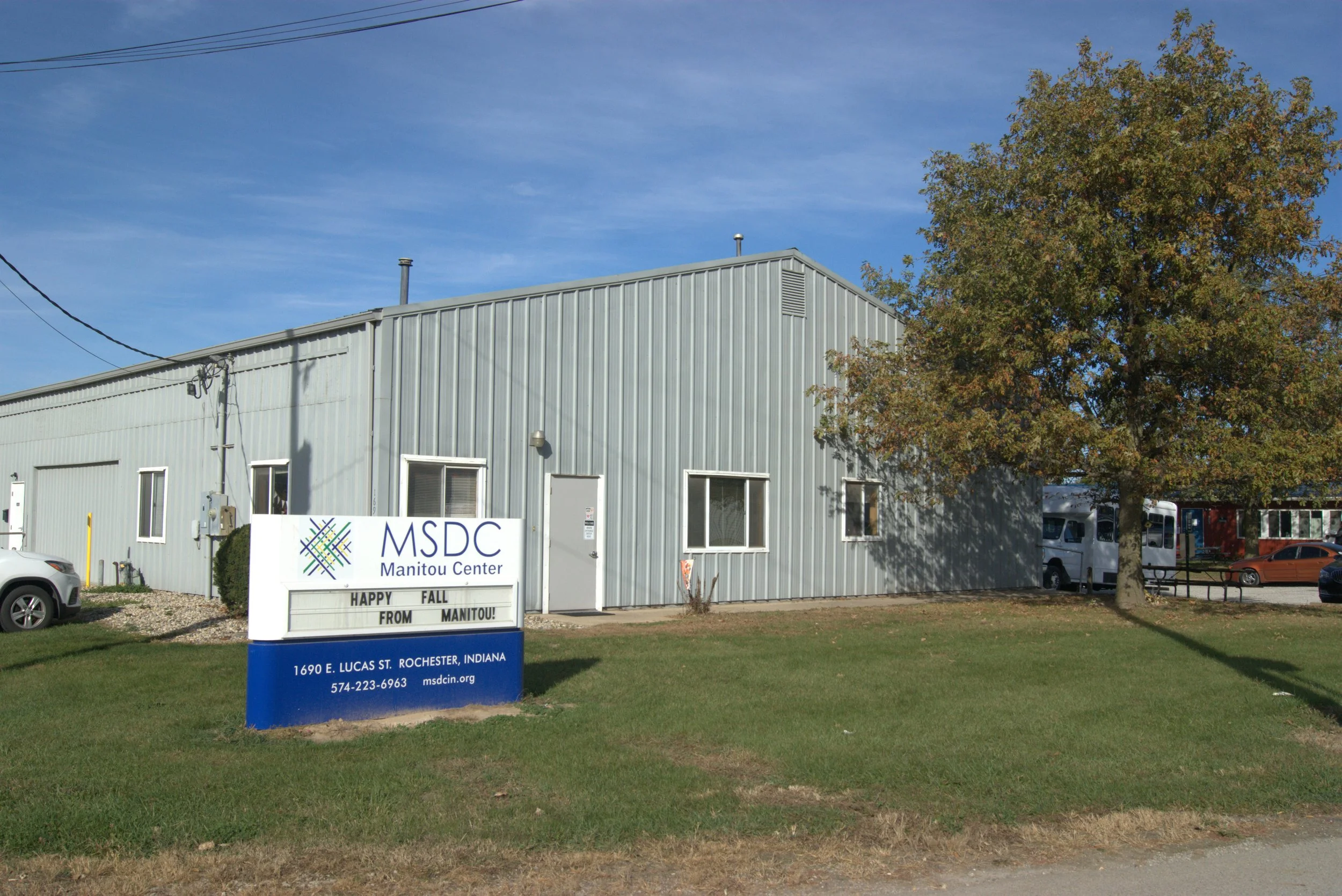 A grey metal building with windows and a door, a sign for MSDC Manitou Center, a tree, some parked cars, a grassy area, and a clear blue sky.