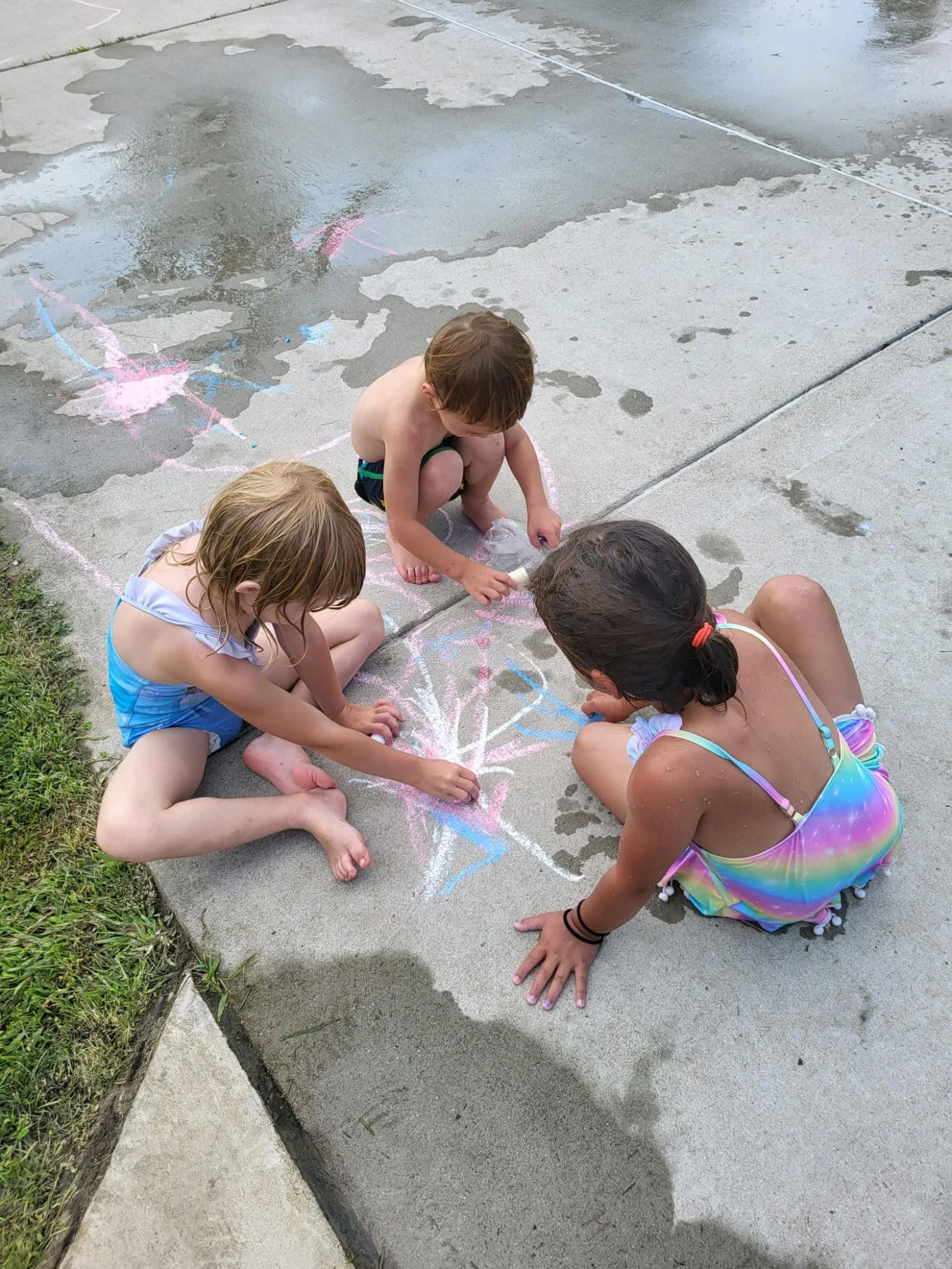 Three children sitting and kneeling on a wet concrete sidewalk, drawing with colorful chalk, with water puddles and grass nearby.