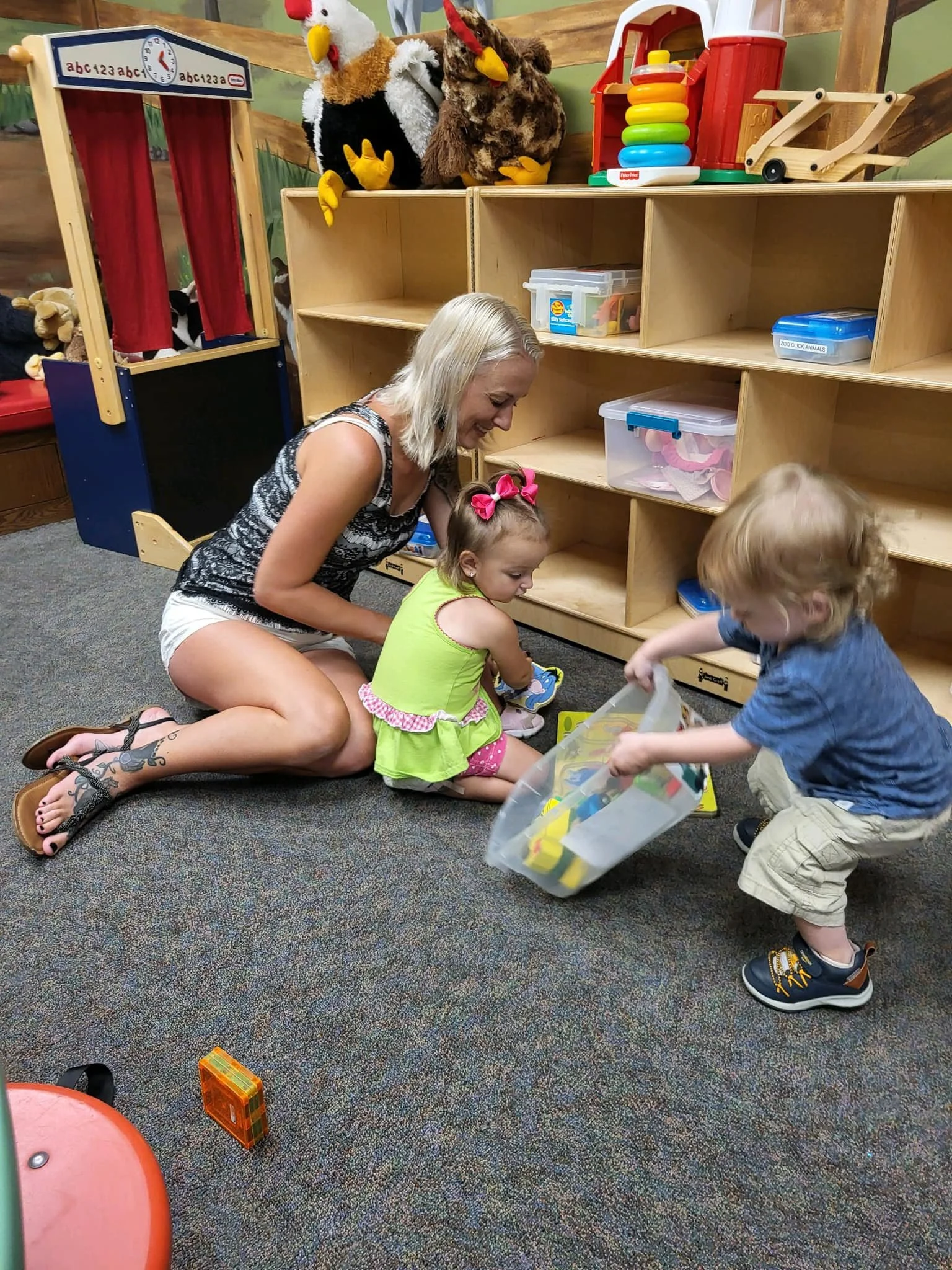 A woman and two children playing with toy building blocks on a colorful carpet in a playroom. The woman is sitting on the floor, smiling, while the children are bending down to pick up or place the blocks. Shelves with toys and stuffed animals are visible in the background, including puppet characters and a stacking ring toy.