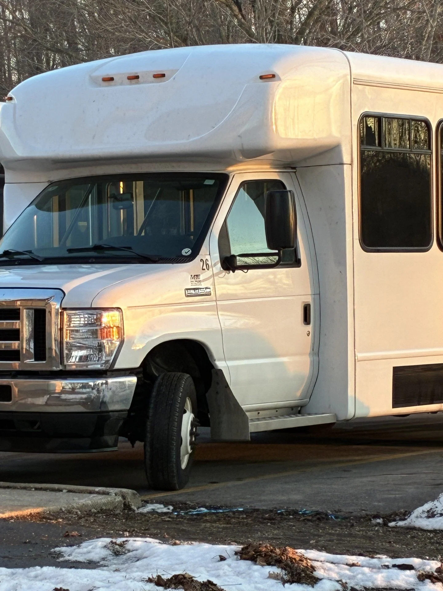 White step van parked on asphalt with snow and leaves on the ground, background of trees and twilight sky.
