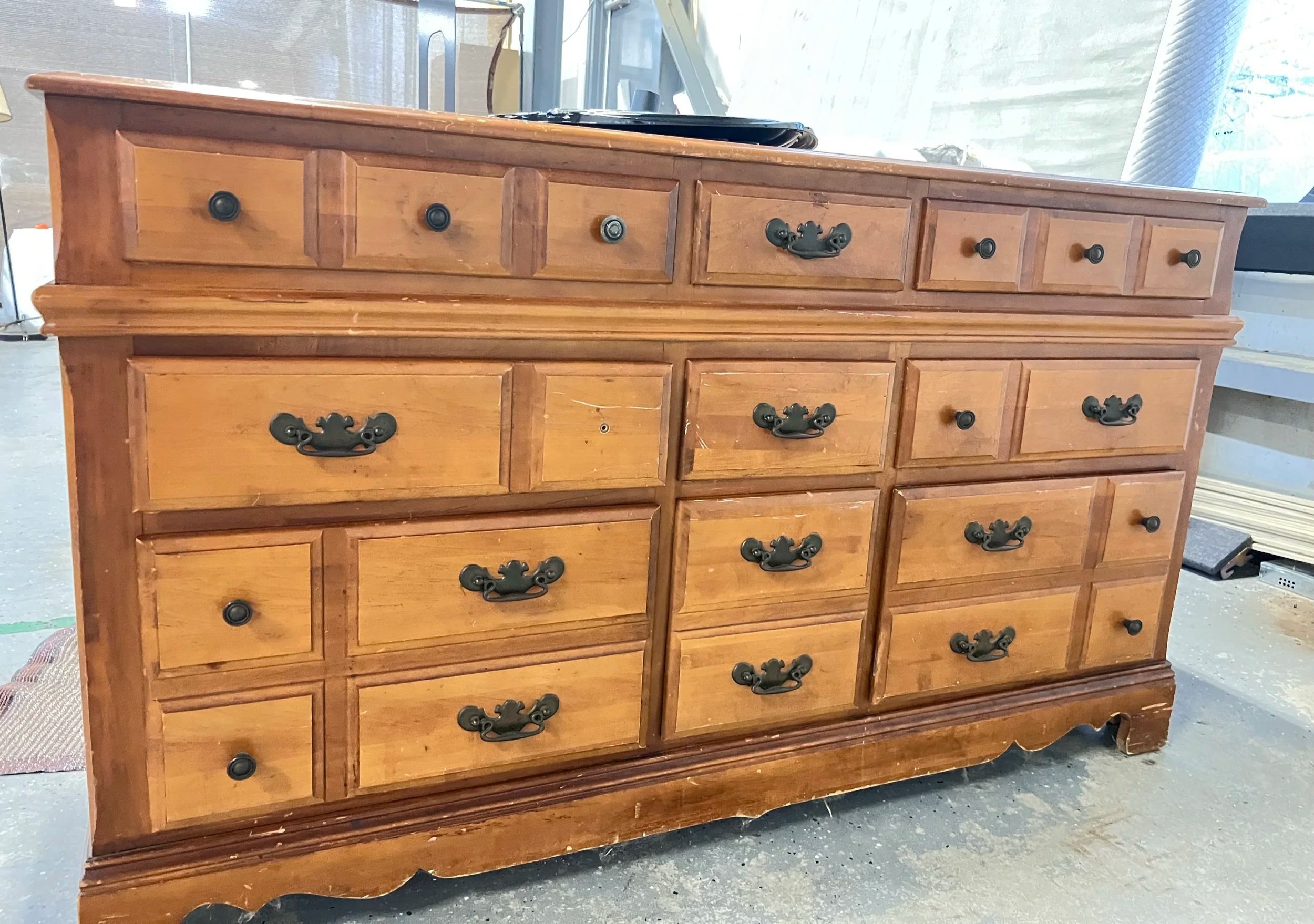A large wooden dresser with multiple drawers, featuring ornate black handles, situated in a room with concrete flooring and a sliding glass door in the background.