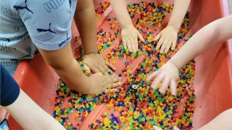 Children's hands playing in a bin filled with colorful plastic gumballs at an arcade or toy store.