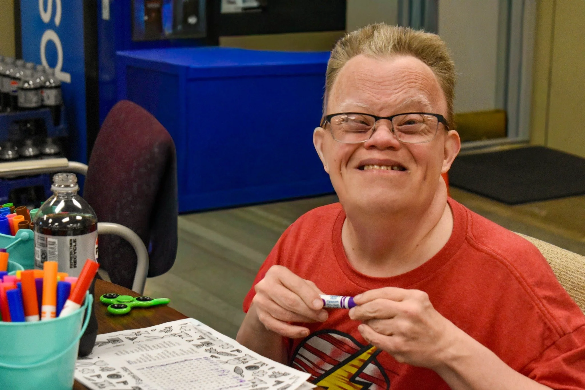 A smiling man with glasses and a red t-shirt sitting at a table with art supplies and paper, holding a glue stick.
