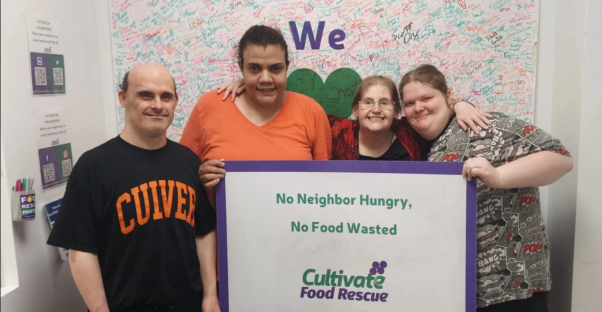 Four people holding a sign that reads 'No Neighbor Hungry, No Food Wasted' in front of a wall covered in signatures and a large green heart. One person wears a black shirt with 'Culver' written in orange, and another wears a shirt with comic book style graphics.