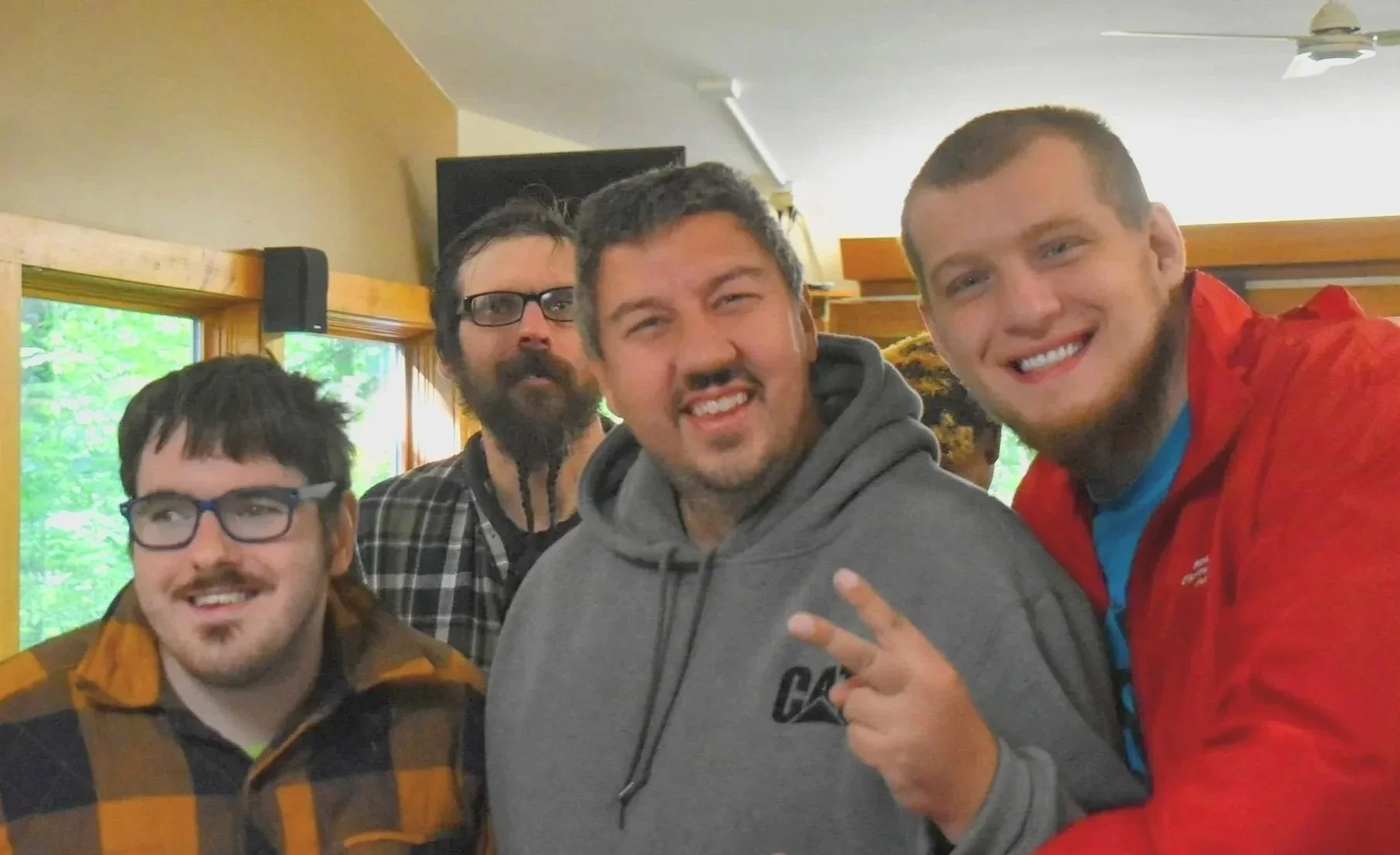 Group of five men indoors, smiling and posing for a photo. They are casually dressed, with one making a peace sign, and a window with greenery visible in the background.