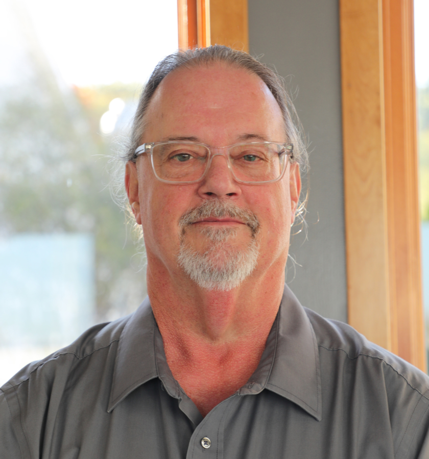A middle-aged man with glasses and a goatee, wearing a gray shirt, standing indoors near a window with wooden trim, with trees visible outside.
