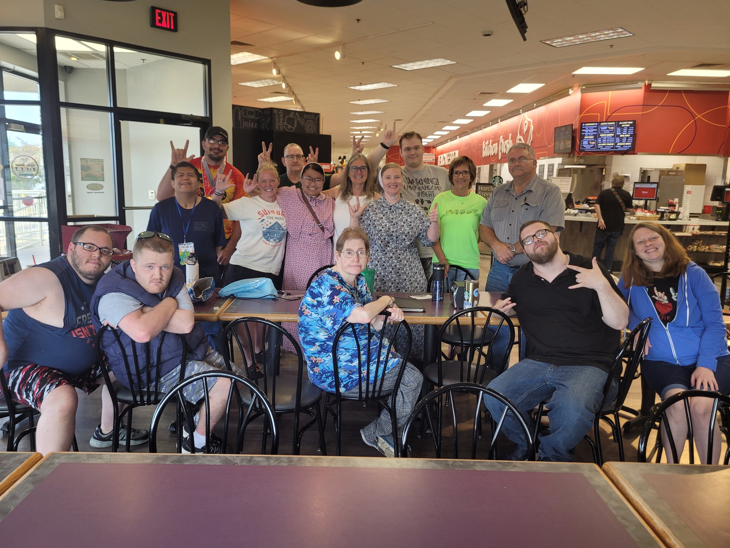 Group of fifteen people gathered around a table in a cafe or restaurant, some standing behind and some sitting in front, smiling and making hand gestures for the photo.
