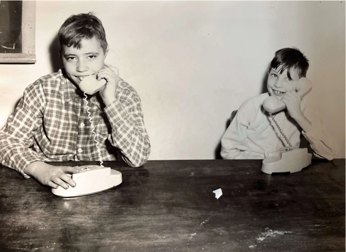 Black and white photo of two young boys sitting at a table, each talking on a landline telephone.