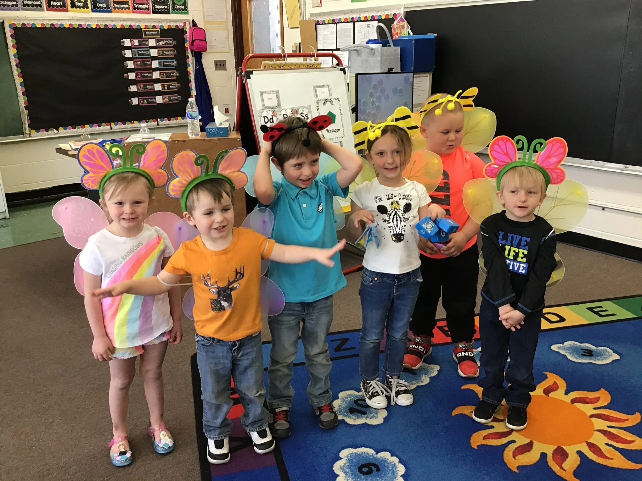 Children in a classroom dressed in colorful fairy and bee costumes, standing on a vibrant outdoor-themed rug with a sun design.