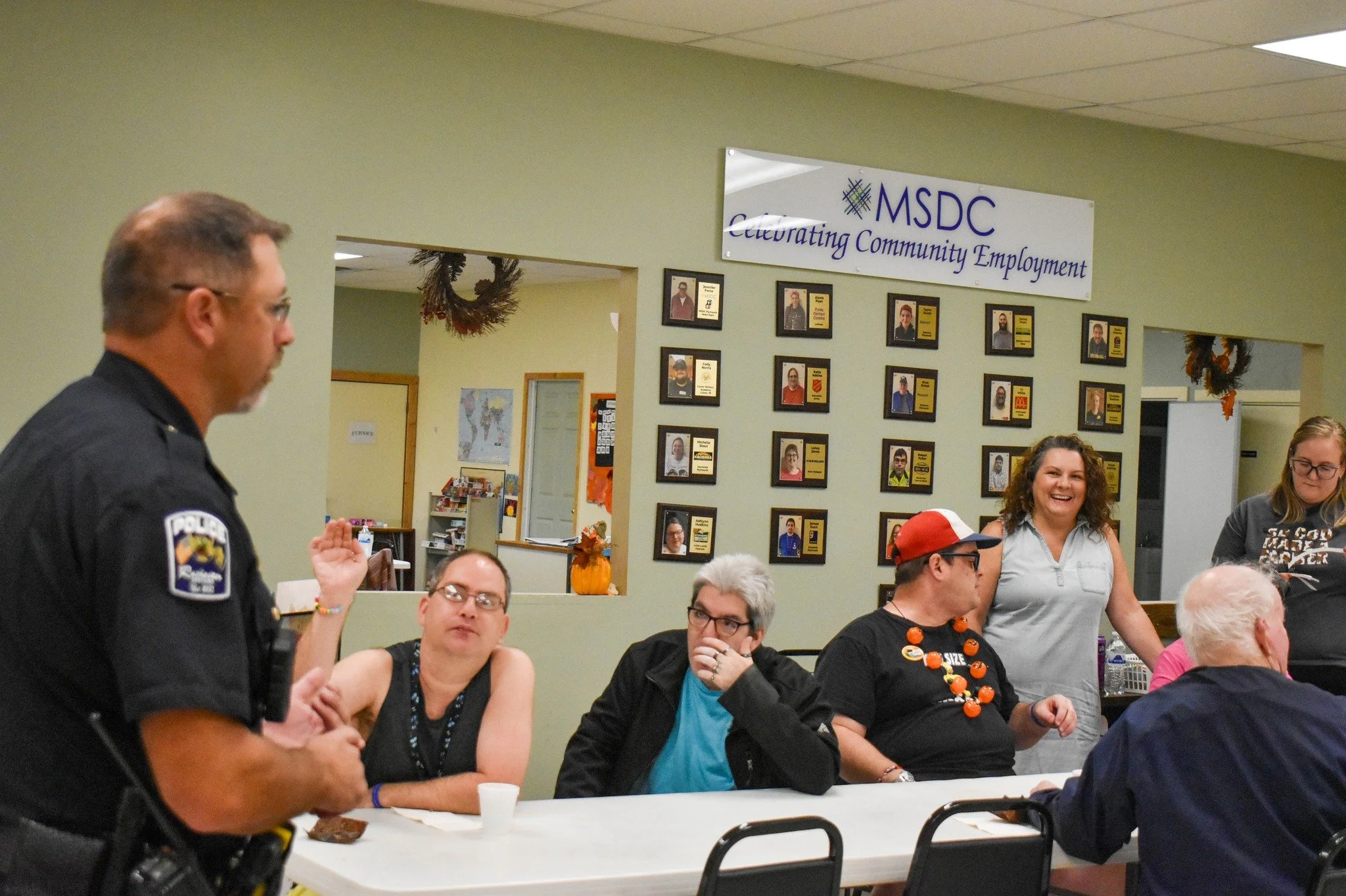 Police officer speaking to a group of adults at a community conversation event, with a wall display of framed photos and awards in the background.
