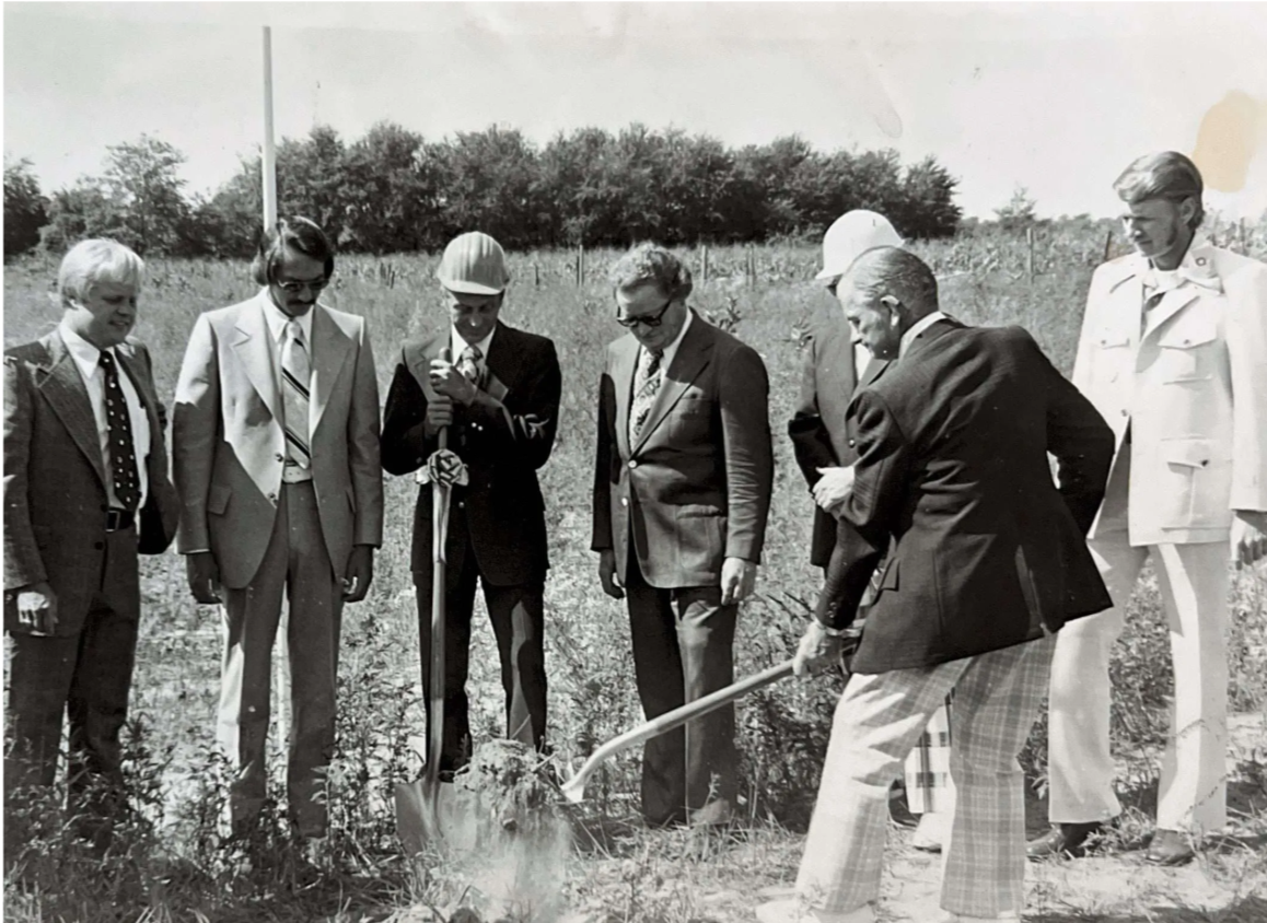 Group of adults watching a person with a shovel plant a tree in a grassy outdoor area.