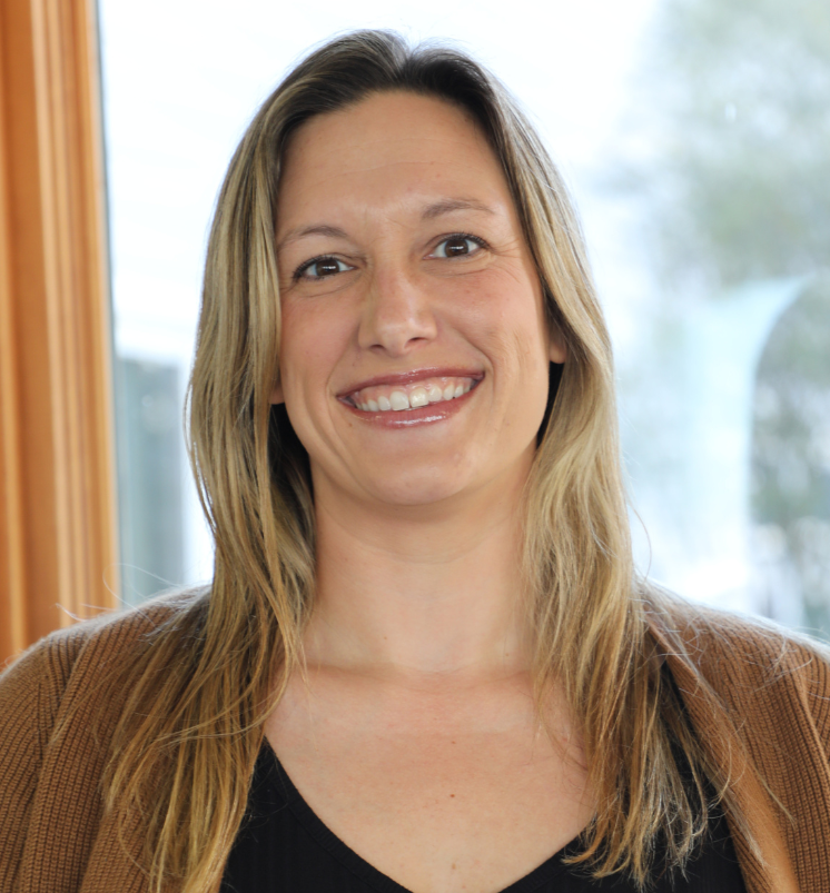 A woman with long blond hair, smiling, wearing a brown jacket and black top, standing indoors near a window.