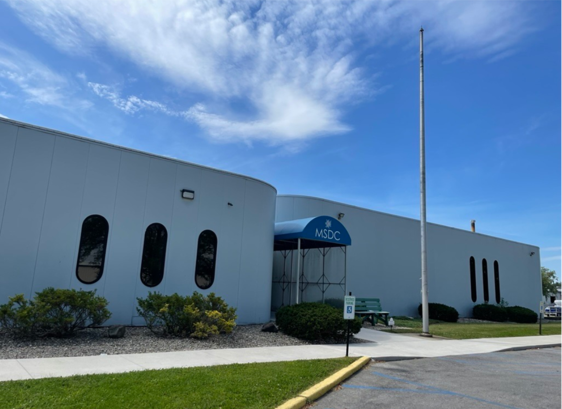 Photo of a modern, white building with rounded walls and narrow oval windows, featuring a sign reading "MSDC," under a bright blue sky with scattered clouds.