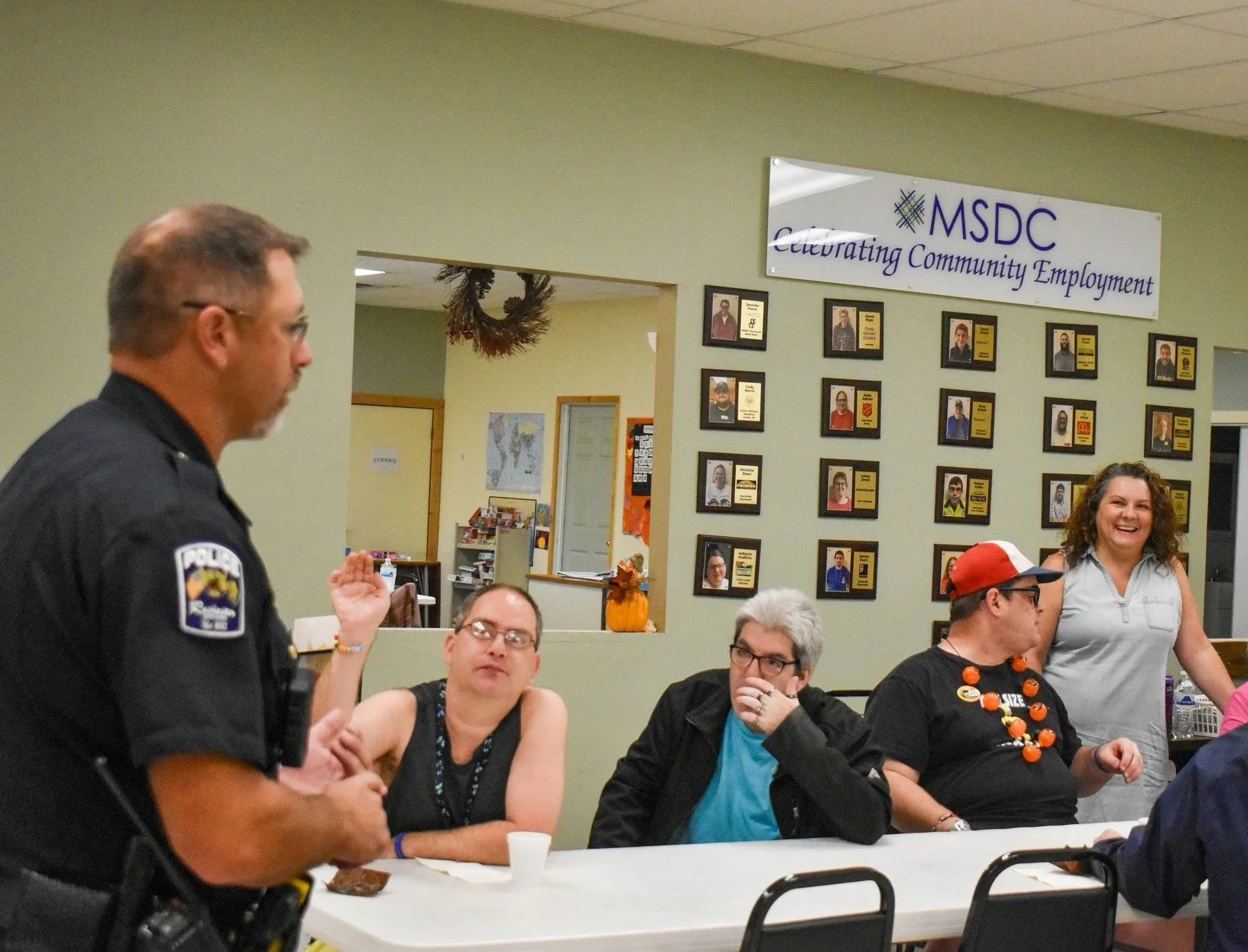 A police officer is speaking to a group of people seated at a table during a community event at MSDC, a community employment organization. The continuation of the event's theme is visible on a wall with framed photographs and a sign that reads 'Celebrating Community Employment'.
