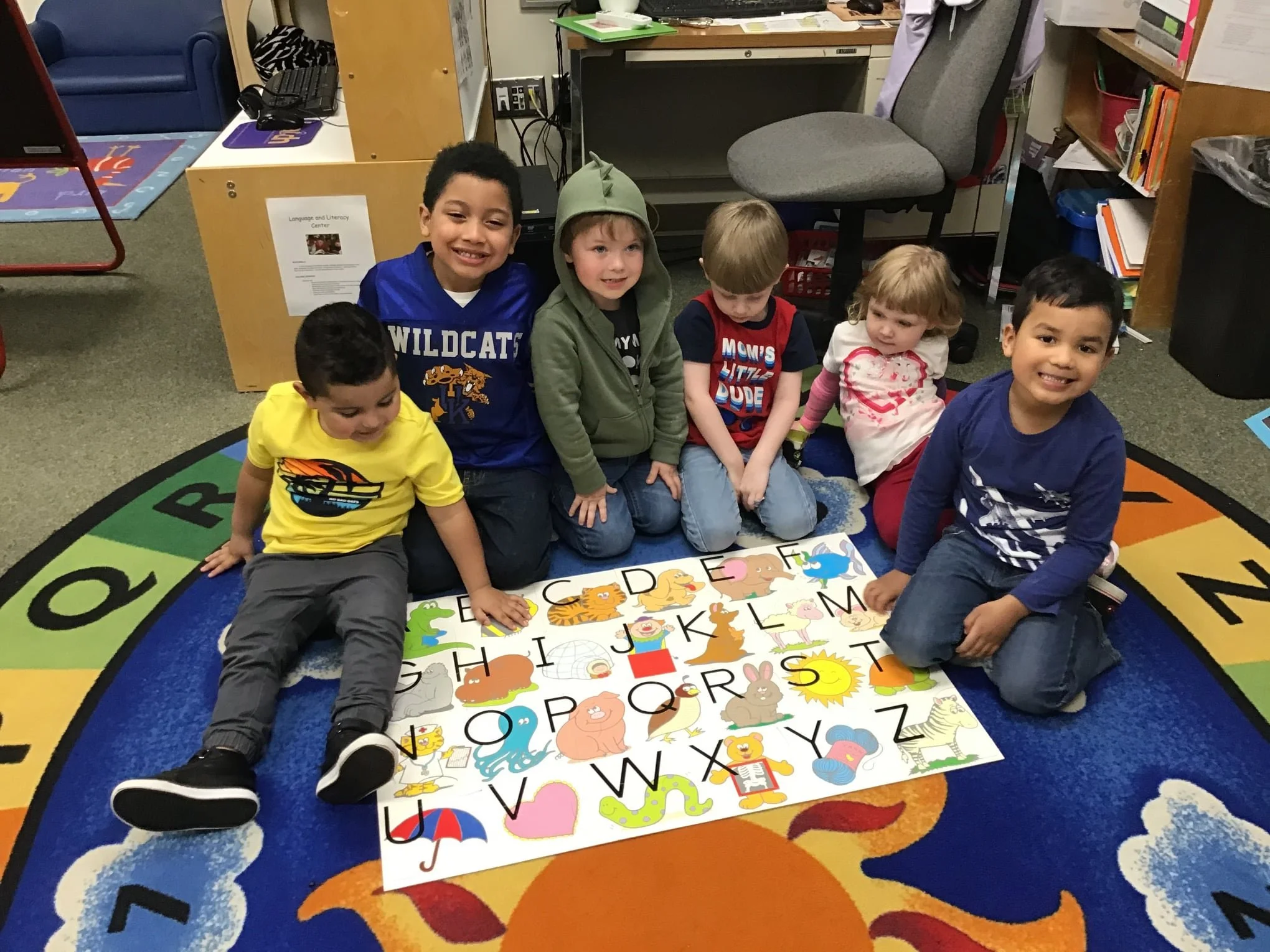 Six children sitting on a colorful alphabet rug in a classroom, smiling at the camera, with a large alphabet puzzle on the floor in front of them.