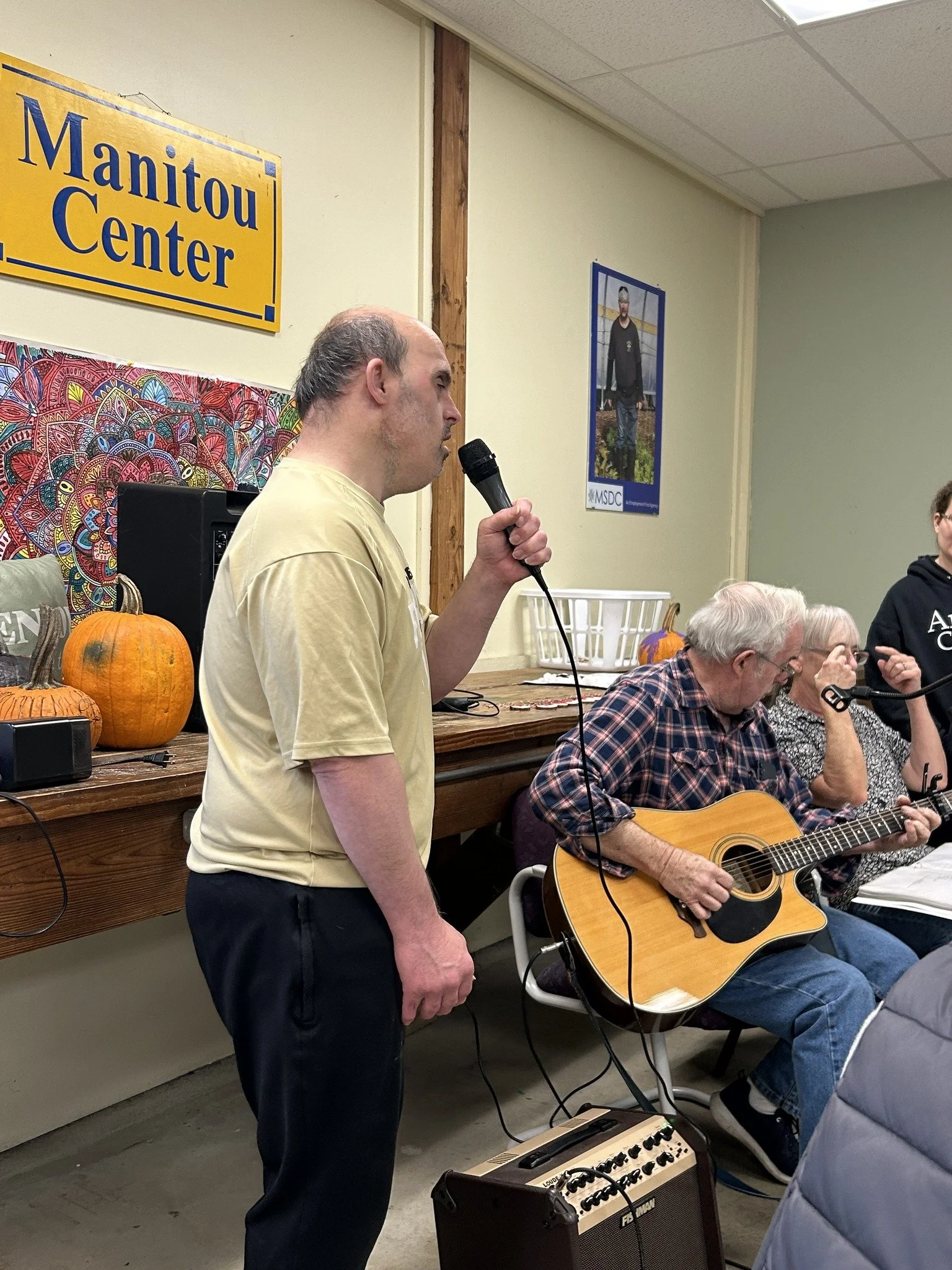 A man holding a microphone sings or speaks, with a man playing an acoustic guitar seated beside him. There are pumpkins on a table behind them, and a 