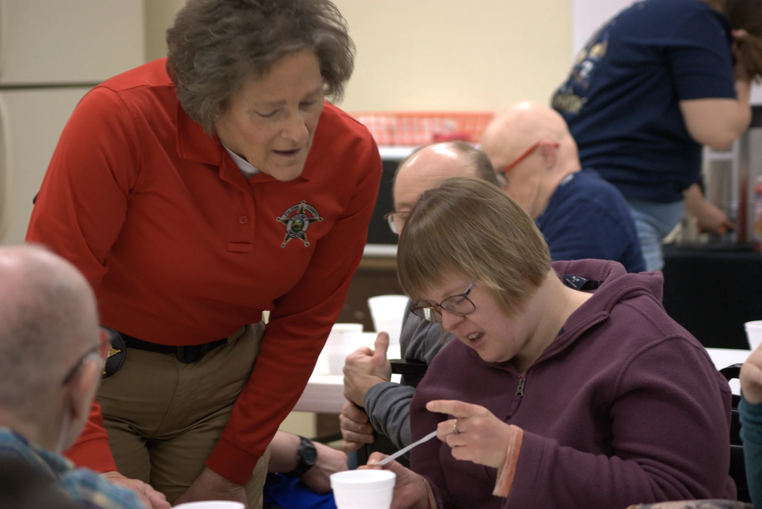 Police officer speaking to a group of adults at a community conversation event, with a wall display of framed photos and awards in the background.