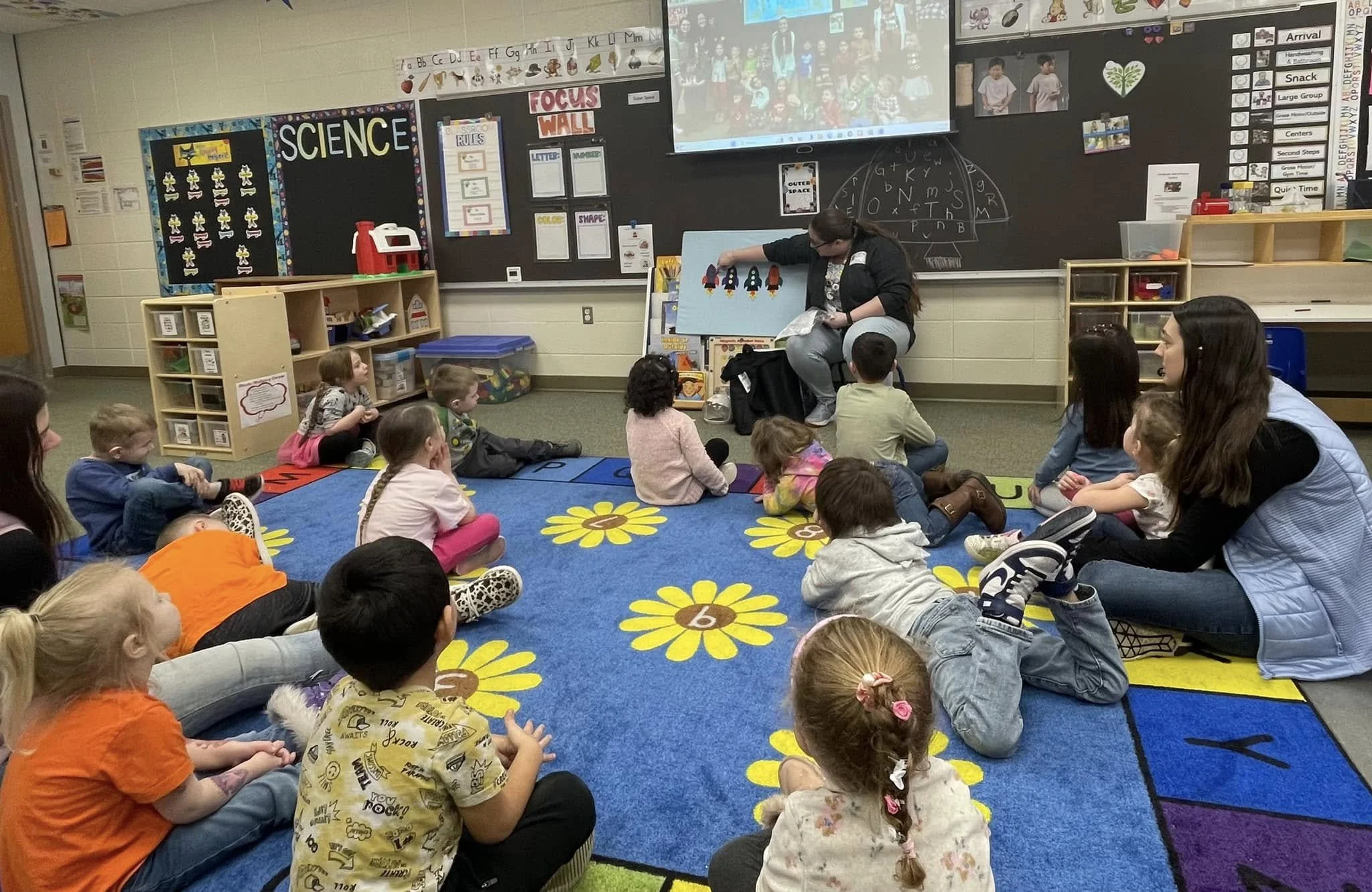 A kindergarten classroom with children sitting on a colorful alphabet carpet, listening to a teacher reading a story and showing an illustration of space rockets. Teachers and children are engaged in a storytime activity.