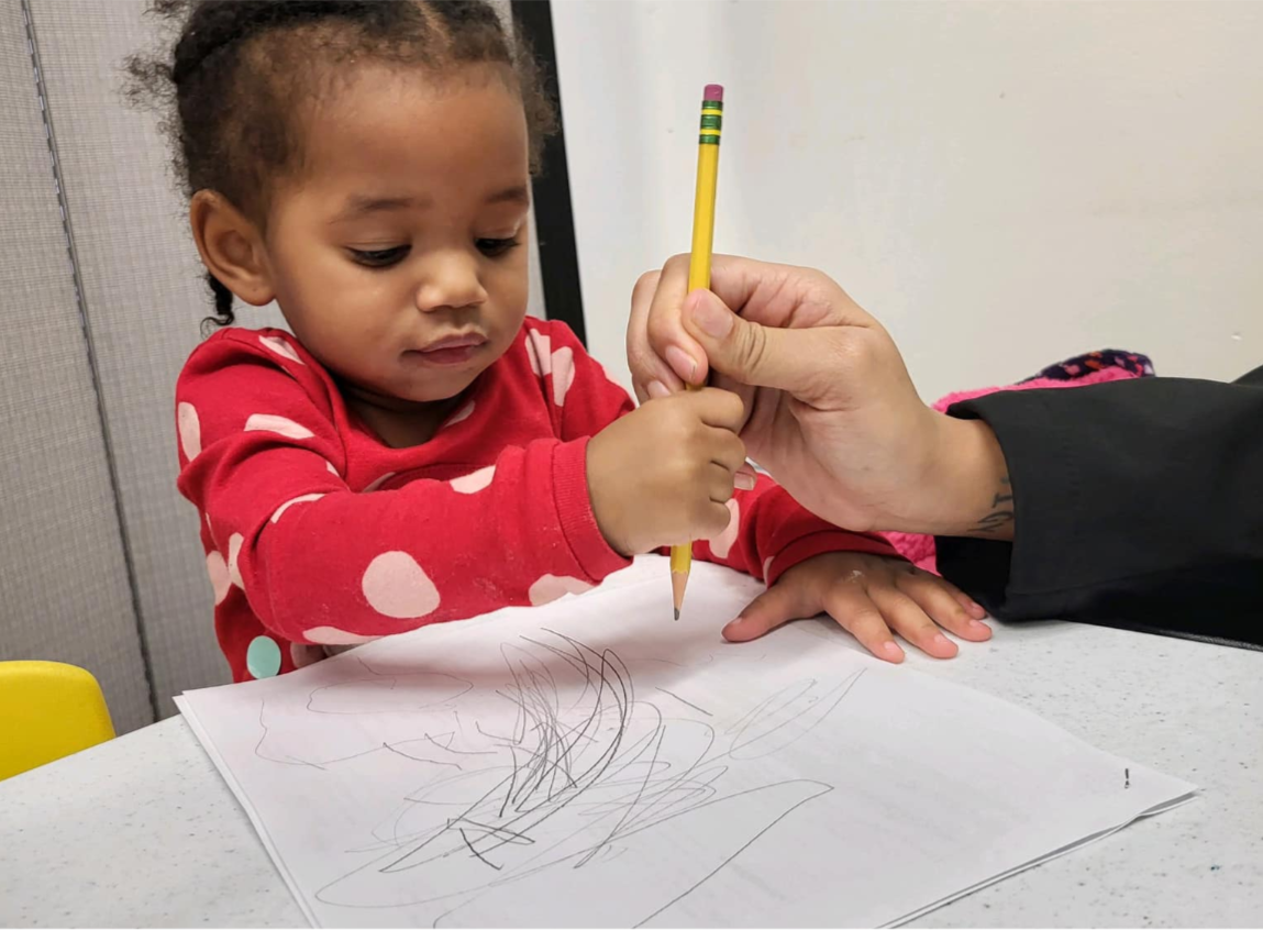 A young girl with curly hair wearing a red shirt with white polka dots draws on paper with a yellow pencil, assisted by an adult's hand.