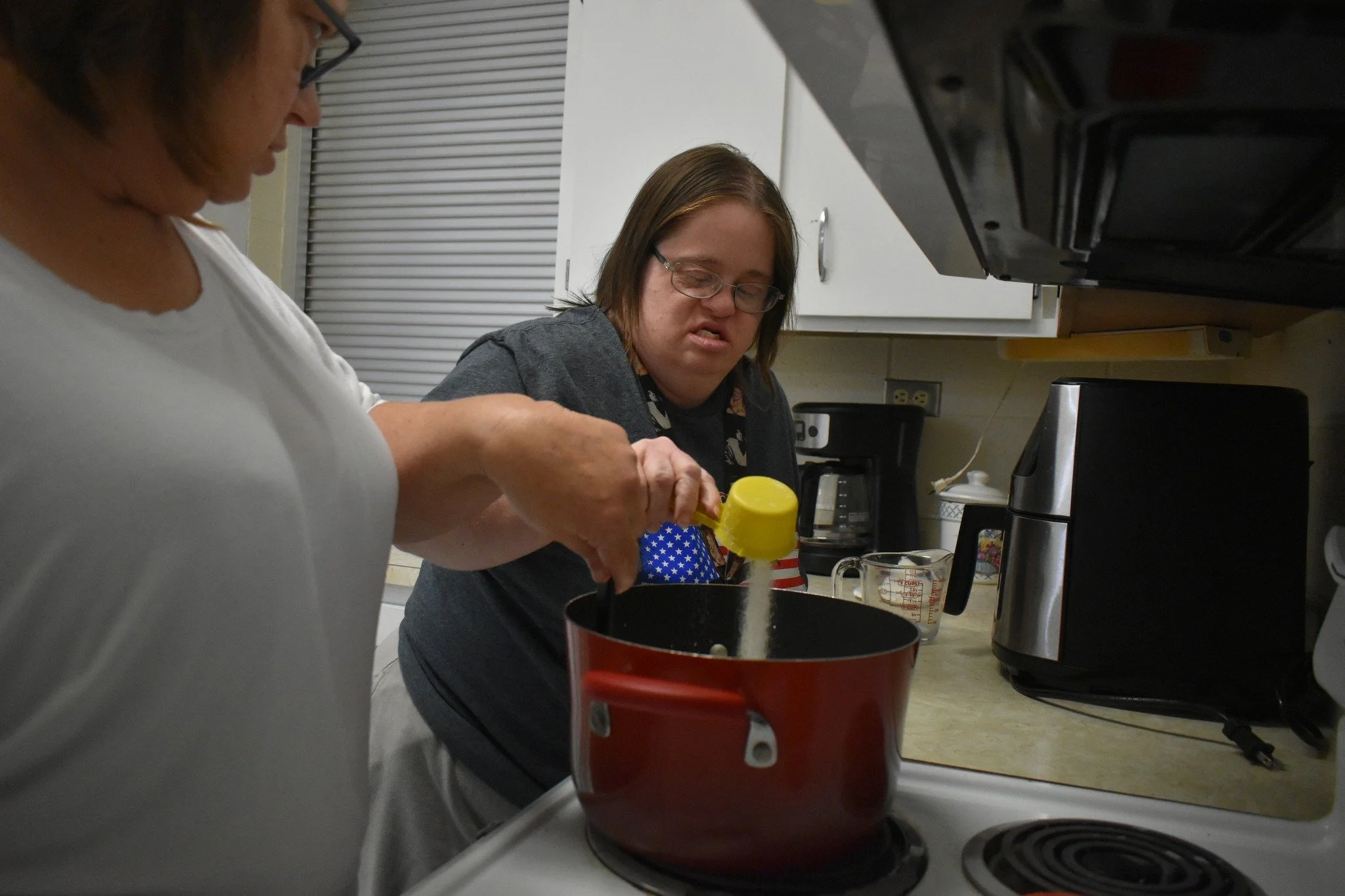 Two women cooking in a kitchen, one adding salt to a pot on the stove, the other observing with a focused expression.