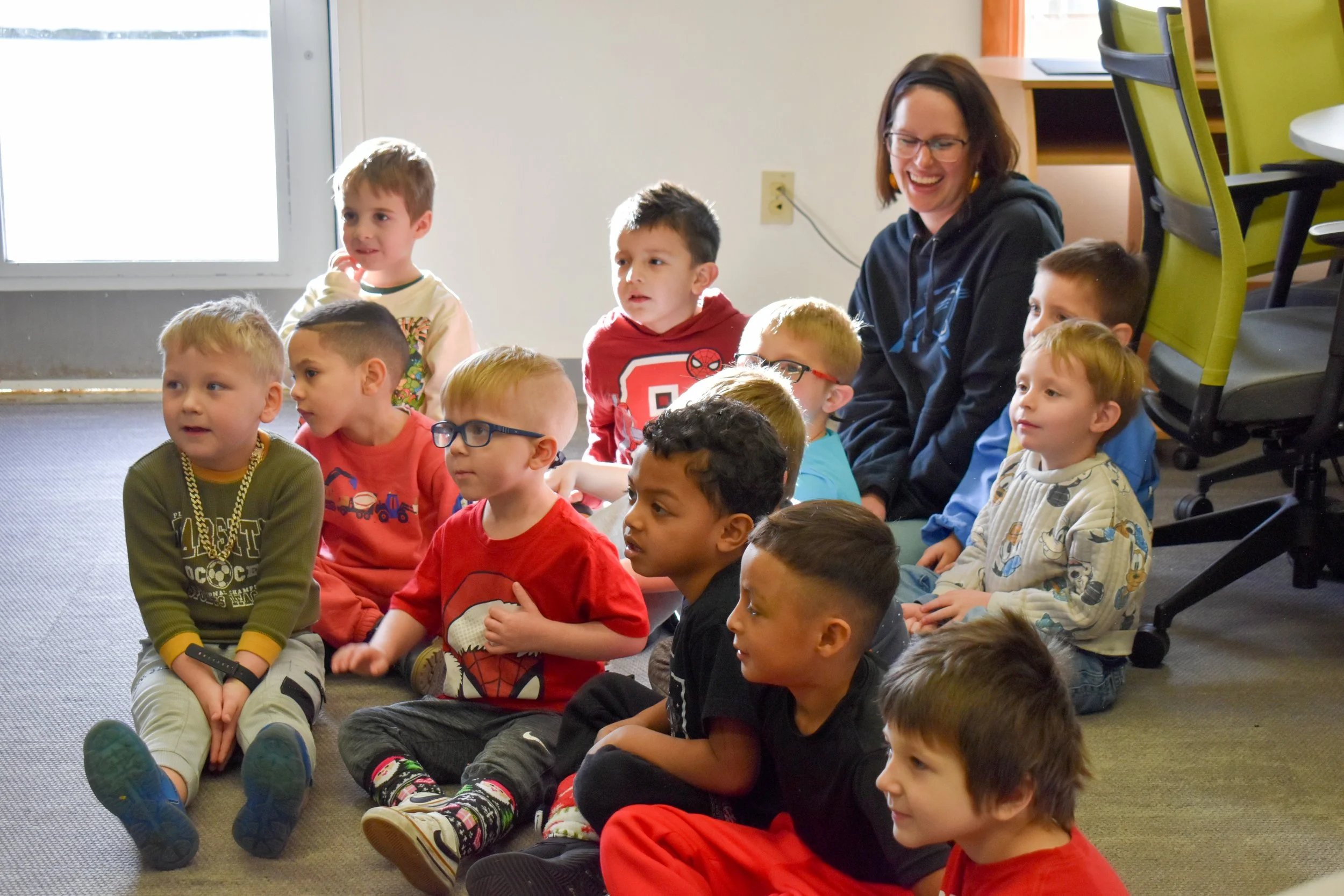A group of young children sitting on the floor indoors with a smiling woman, possibly a teacher, in a classroom setting.