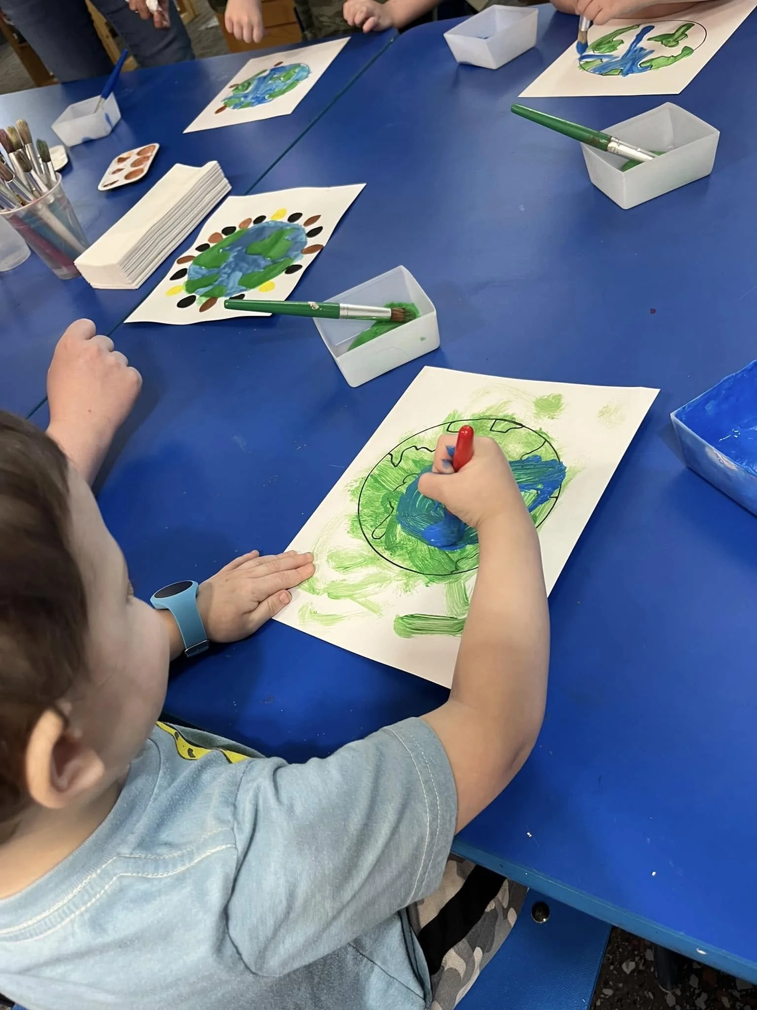 Children painting and drawing pictures of Earth on white paper with green, blue, and black markers and paints, sitting around a blue table.