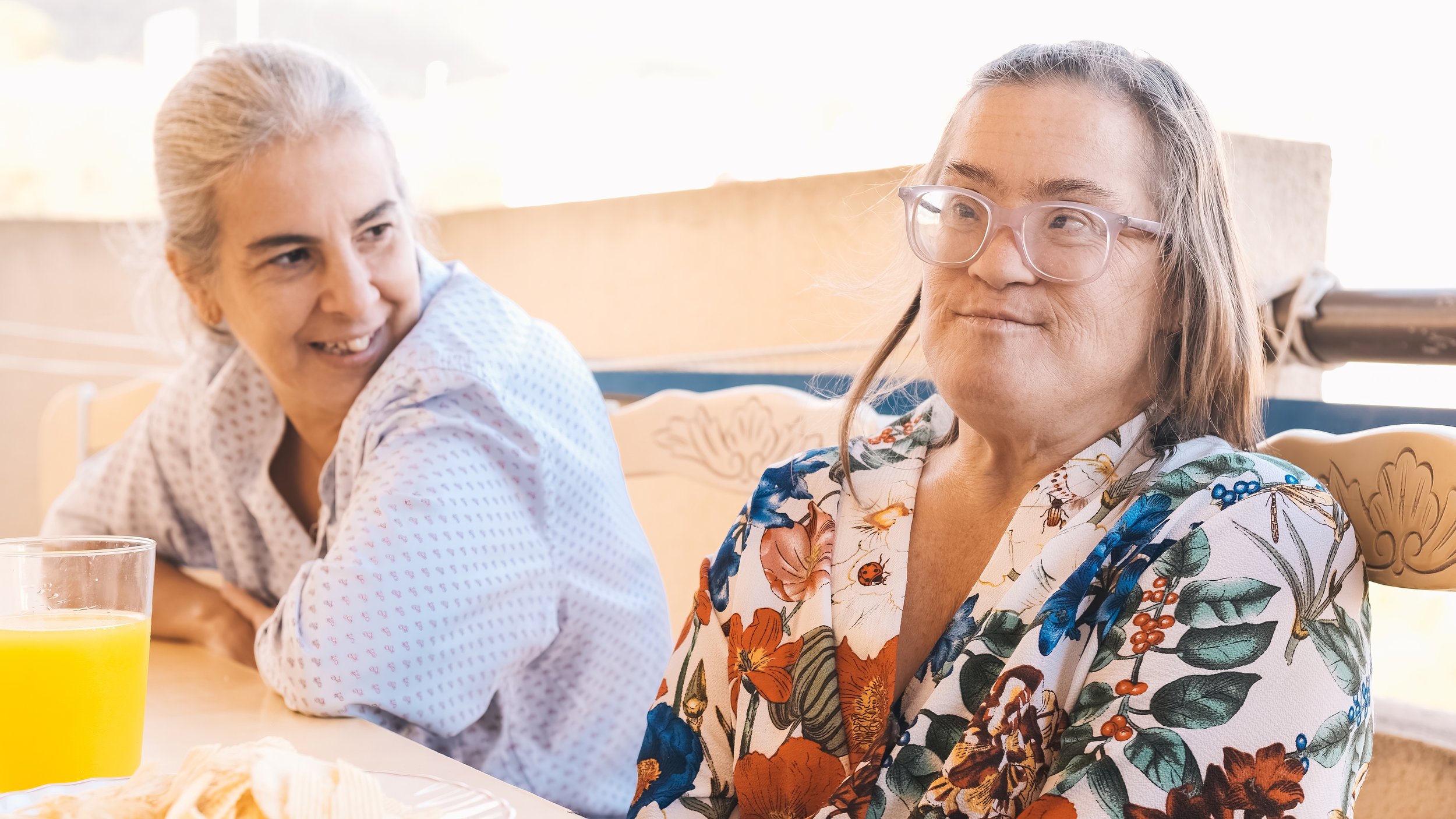 Two women sitting at a table, one older with gray hair in a bun, smiling, wearing a white blouse with pink pattern; the younger woman wearing glasses and a floral blouse, looking to the side with a slight smile.