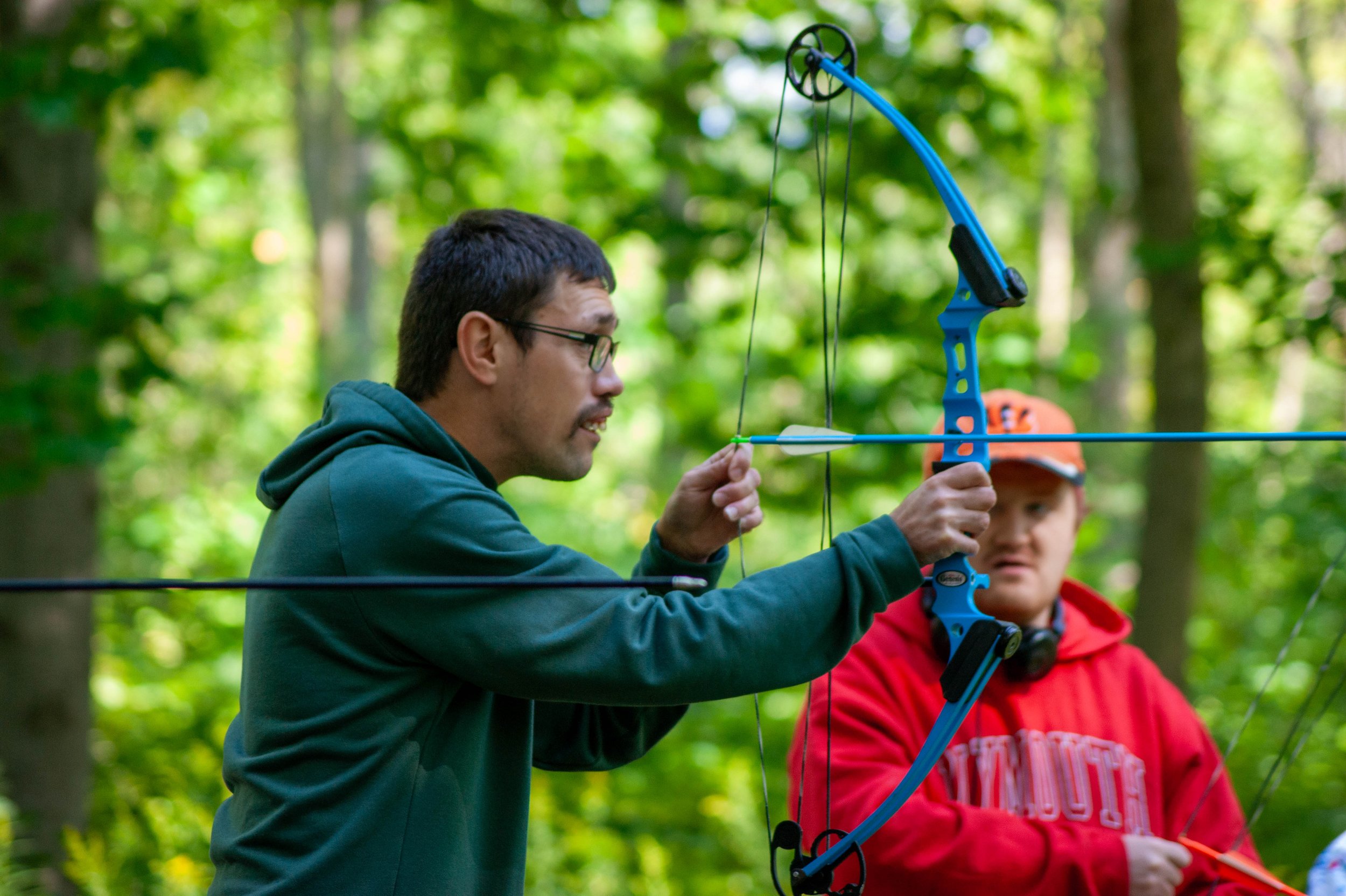 Two people in a forest, one man aiming a blue bow and arrow while another person in a red jacket watches.