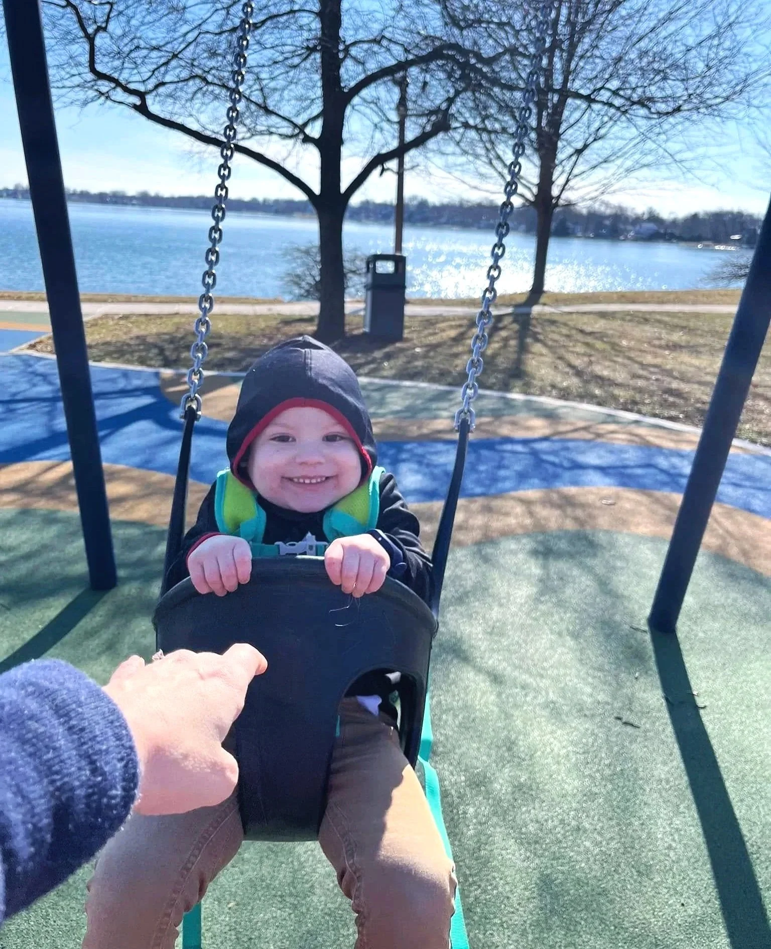 A young boy smiling and sitting on a swing at a park near a lake, with trees and a walking path in the background on a sunny day.