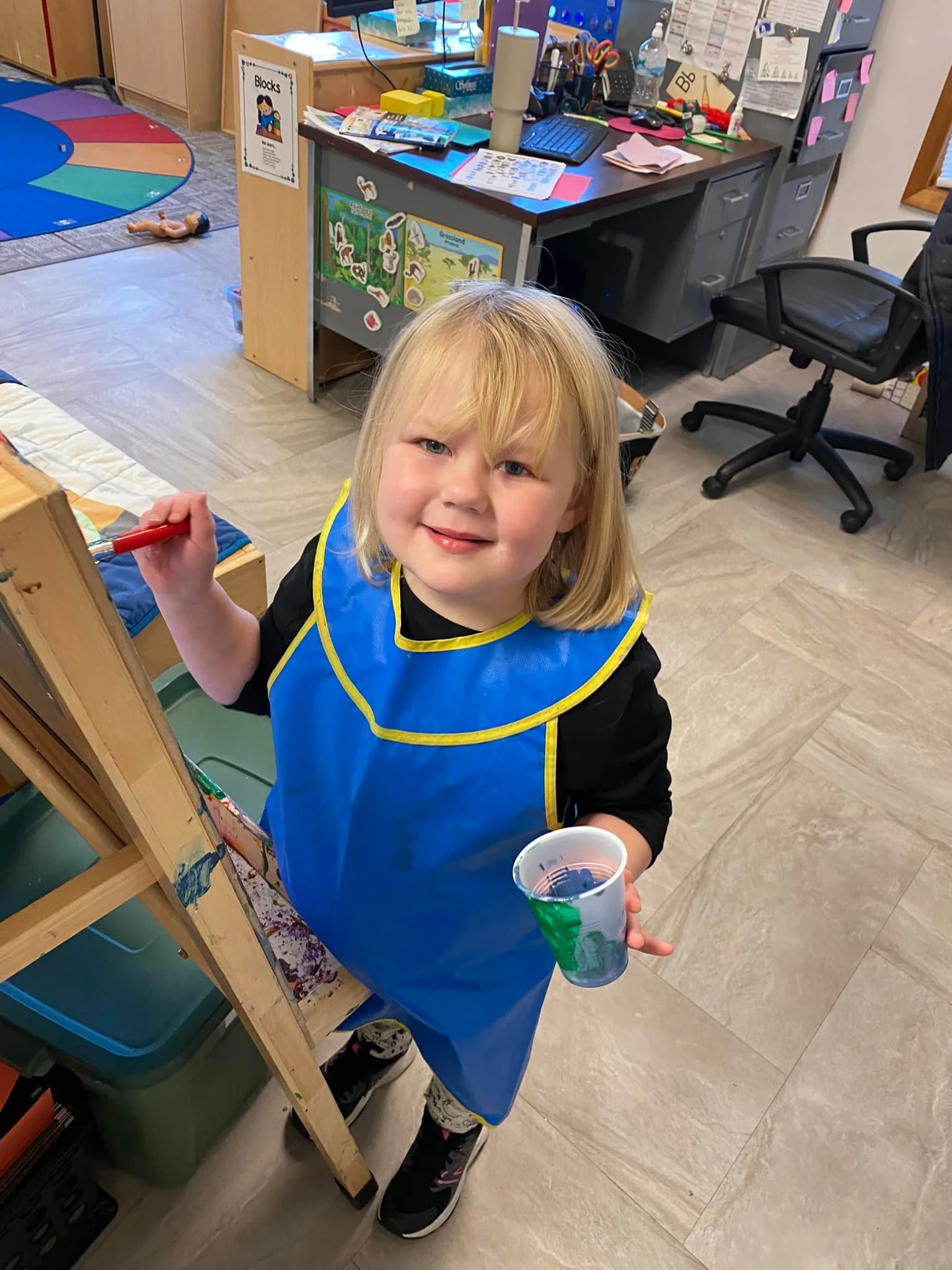 Young girl with blonde hair wearing a blue apron, holding a cup of paint and a paintbrush, smiling in a classroom or art room.