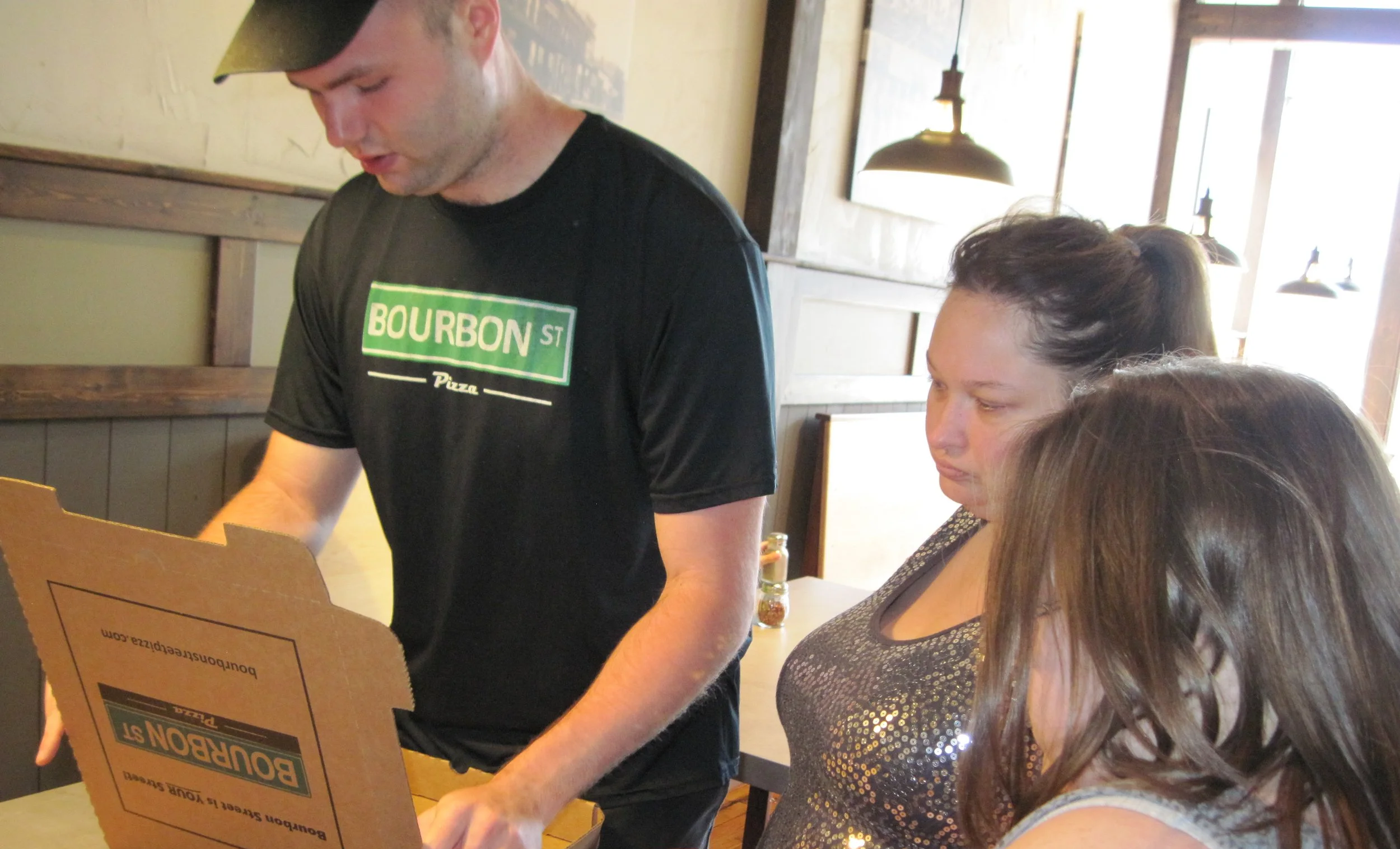 A man wearing a black BOURBON ST pizza t-shirt and a cap appears to be preparing or serving pizza inside a rustic restaurant, with two women watching.