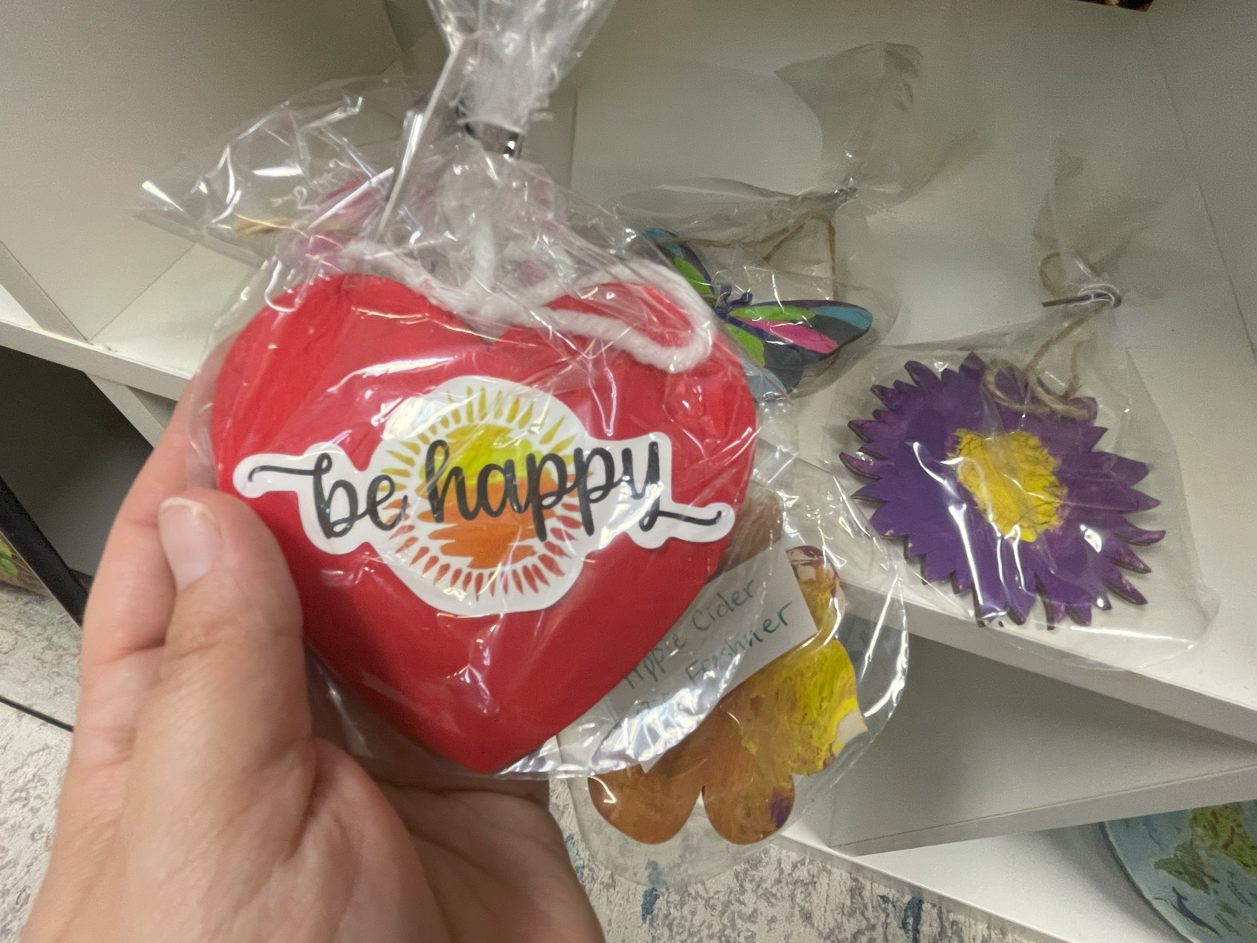 A hand holding a red decorative apple with a sticker that says 'be happy.' Behind are flower-themed decorations in plastic packaging on a white shelf.