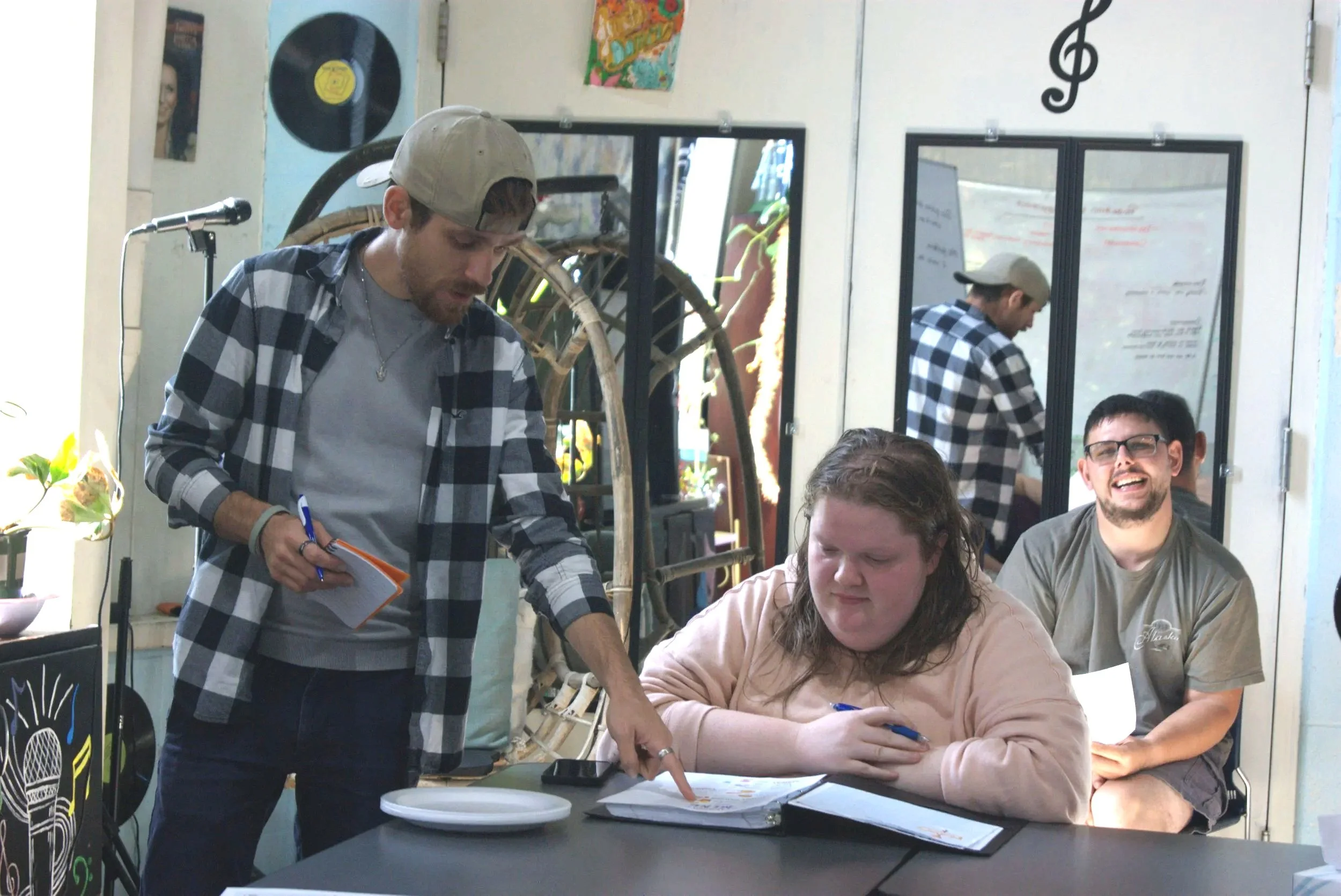 A group of people in a casual setting: a man with a plaid shirt and cap pointing at a notebook, a woman with wet hair looking at the table, a man with glasses laughing, and another person in the background. There are mirrors, musical notes, and decorations on the walls.