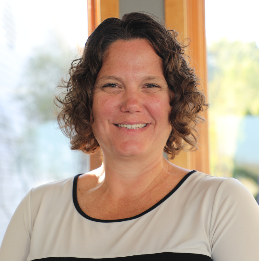 Smiling woman with short curly brown hair, wearing a white top with black trim, indoors with a window showing trees in the background.