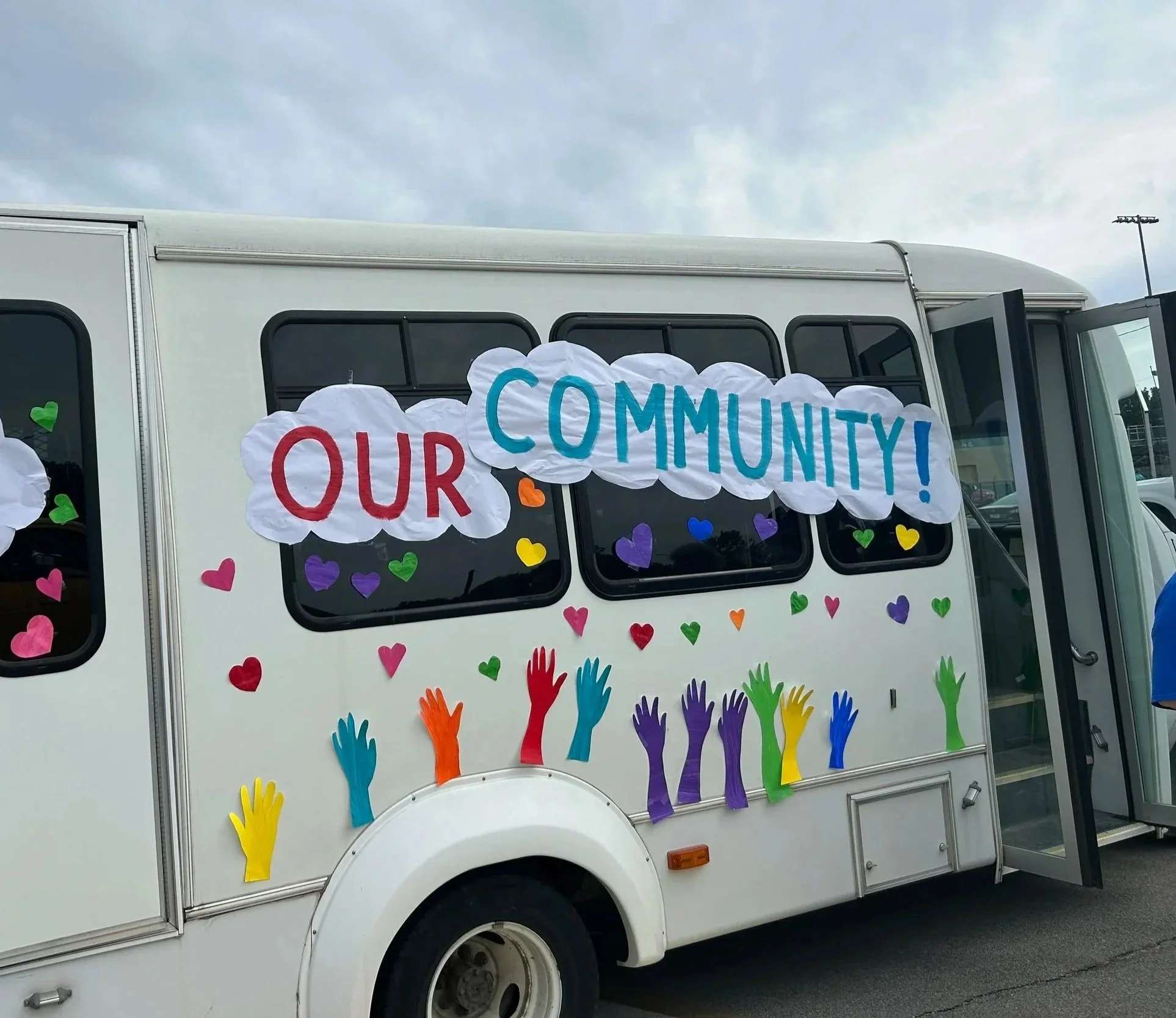 A white bus decorated with colorful paper hearts and cutouts of raised hands, featuring a sign that says 'Our Community!' in red, blue, and teal letters.