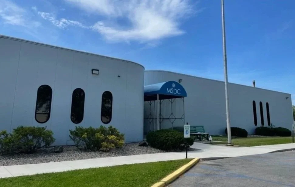 Exterior view of a white commercial building with a blue awning labeled 'MSDC,' surrounded by green shrubs, a grassy lawn, and a parking lot with a handicapped parking sign, under a partly cloudy sky.
