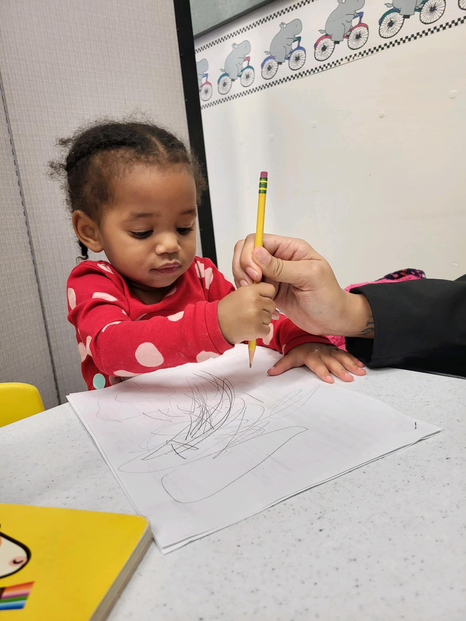 A young girl in a red polka-dot shirt draws on a piece of paper with a yellow pencil. An adult hand holds the pencil, helping her. The girl has short curly hair and is sitting at a white table. There is a yellow book on the table and a whiteboard in the background with a cartoon bear riding a bicycle.