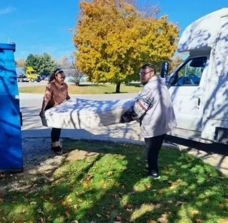 Two people loading a mattress into a white truckk with fall-colored trees.