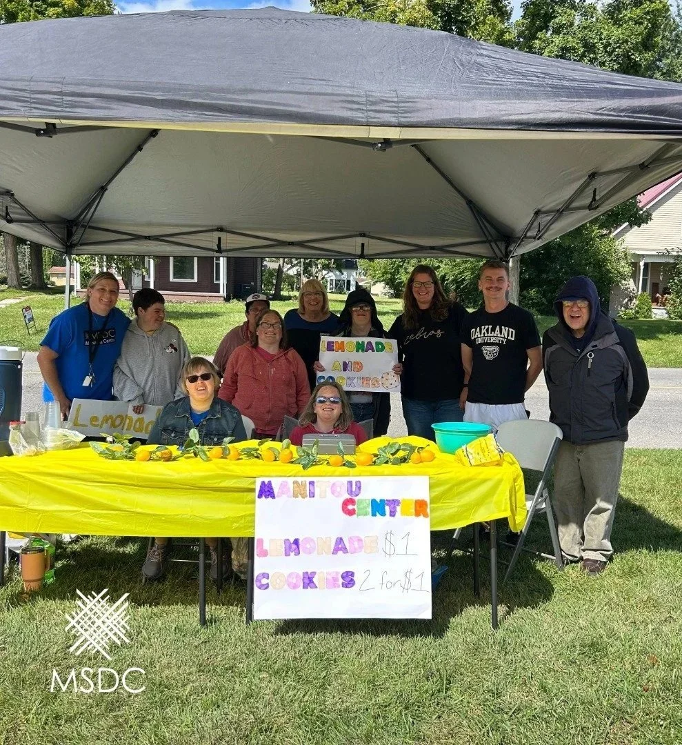 A group of people standing under a black canopy behind a table decorated with lemons and green leaves. The table has signs advertising lemonade and cookies for sale. Some people are holding signs, and there is a sign in front of the table with prices for the lemonade and cookies.
