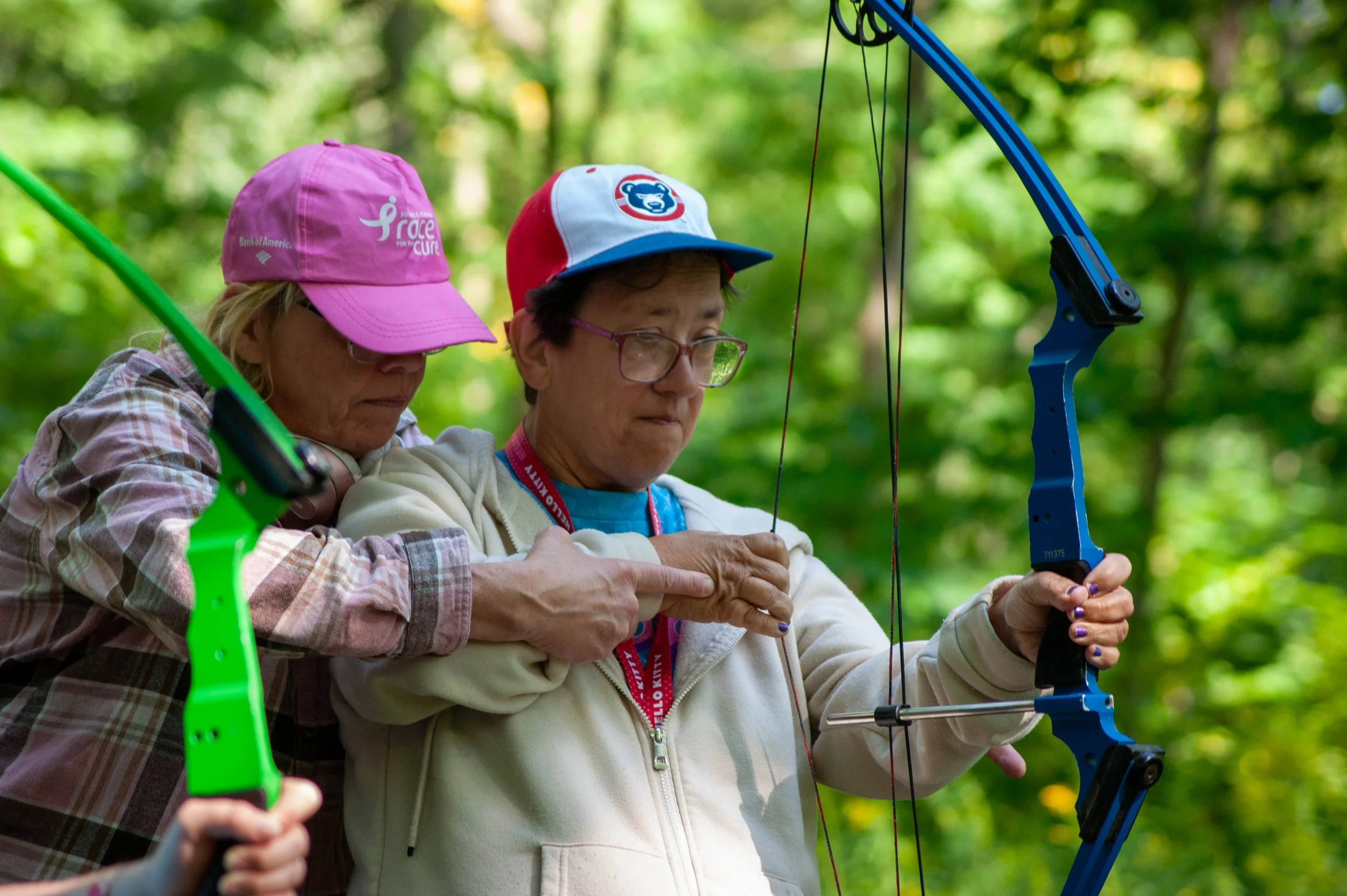 Two women outdoors in a wooded area, one helping the other with archery; one woman is holding a blue bow, and the other appears to be instructing or guiding her.