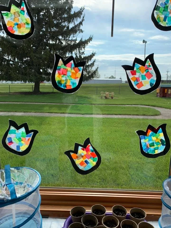 Colorful paper craft tulip shapes with mosaic pattern, hanging on a window, with a grassy outdoor scene in the background.